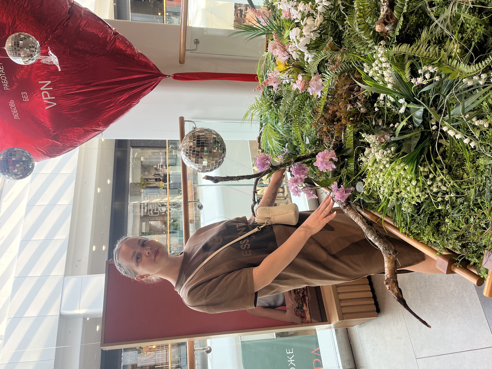 A sideways, medium shot shows a fair-skinned young woman in an Izhevsk, Russia shopping mall. She stands next to a large flower display with pink and white flowers and green plants. She wears a brown t-shirt with a design on the front, and a small, beige purse across her shoulder. She is touching a branch in the display. Above and behind her are decorations including a red, teardrop-shaped balloon with the word "VPN" written in Cyrillic script, and two disco balls suspended from the ceiling. In the background, shops are visible through glass windows. The floor is light grey tile.