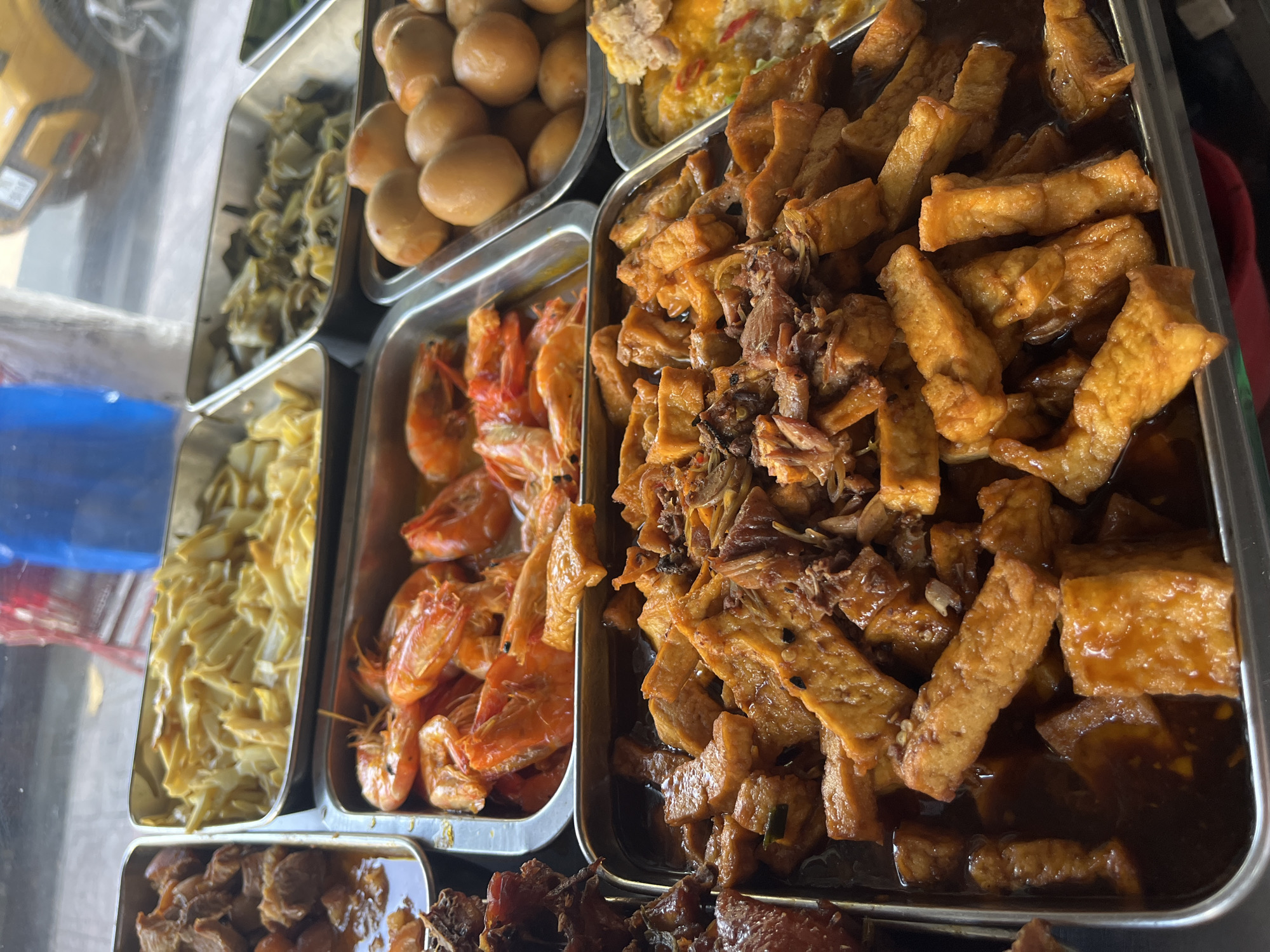 The image shows a selection of dishes in Nha Trang Ward, Vietnam, likely from a food stall or restaurant display. The dishes are arranged in stainless steel trays. Visible are: boiled eggs with browned shells, stir-fried vegetables, cooked shrimp, a dish with fried tofu and meat in a dark sauce, and possibly braised meat. The food appears freshly prepared. A yellow vehicle is visible in the background.