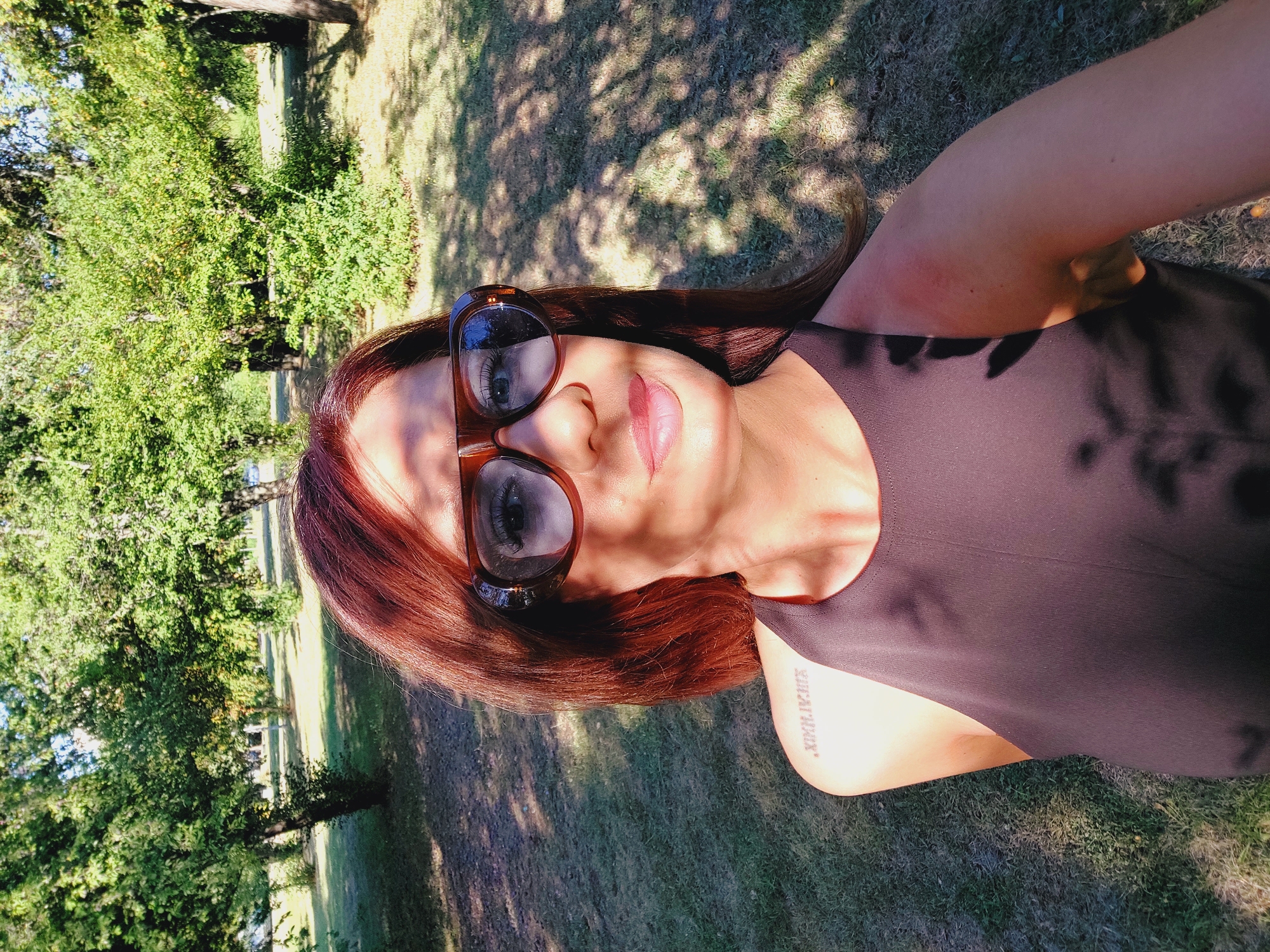 A medium shot captured from a slightly high angle shows a woman with auburn hair and sunglasses posing outdoors in a park. She is looking directly at the camera, and her face is partially illuminated by sunlight, casting shadows from overhead leaves. She is wearing a dark, sleeveless top, and a tattoo in Roman numerals, "XIX.IV.XIX," is visible on her left shoulder. The background consists of a grassy area with scattered trees and dappled sunlight filtering through their foliage. The overall setting suggests a sunny day in a park.