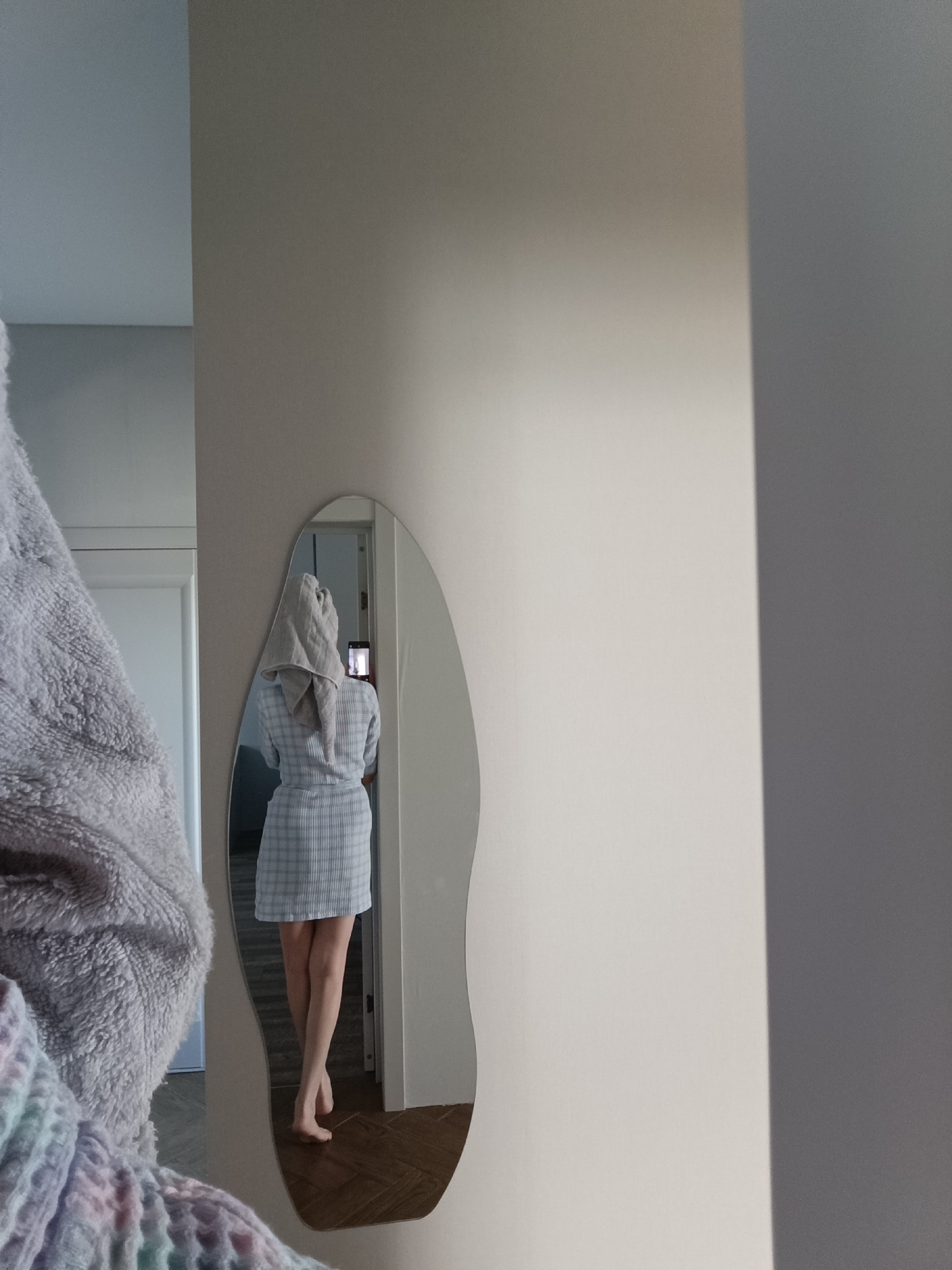 The image displays an indoor scene in Chaikovsky, Russia, featuring a person taking a mirror selfie. A lean individual, seen from behind, is reflected in an irregularly shaped, wavy-edged mirror. The person wears a light blue and white plaid short robe or dress and has a grey towel wrapped turban-style around their head. They are barefoot, standing on a dark brown wooden floor. The individual is holding a smartphone horizontally with both hands at chest level, positioning it to capture their reflection. The phone's screen is illuminated. A white door frame is visible in the reflection to the left. The mirror is mounted on a plain light beige wall. A large, textured, fluffy grey and pastel-colored textile object partially obscures the left foreground of the image. The wall area to the right of the mirror is also light beige, with a distinct vertical shadow line on the far right.