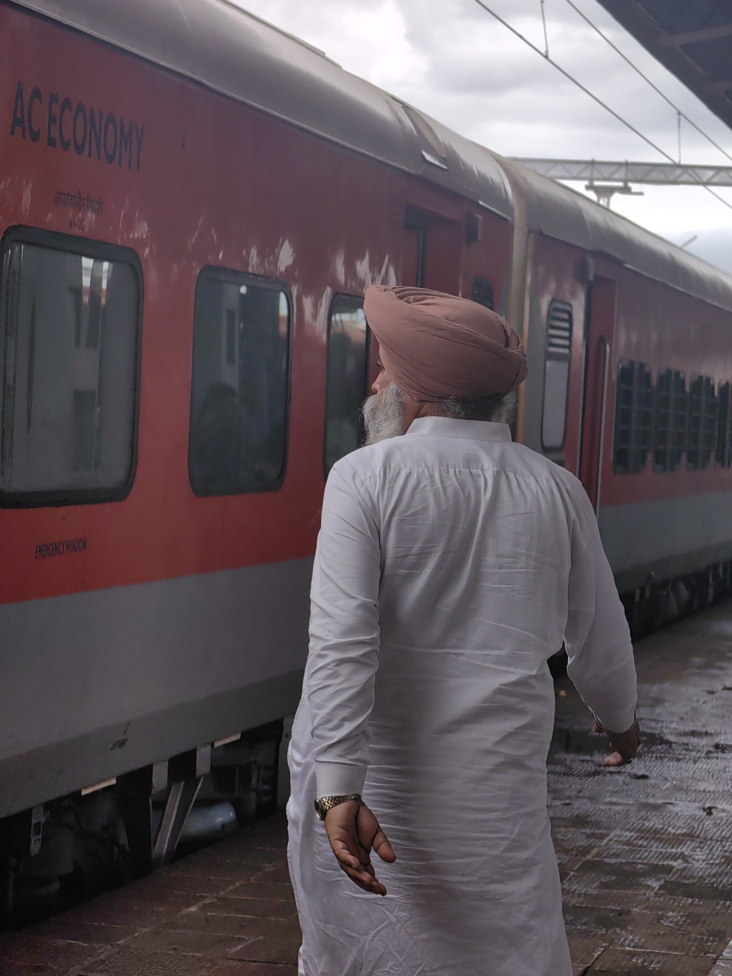 The image depicts a man wearing a turban and a white kurta, standing on a wet train platform in Itarsi, India. He is facing away from the camera, towards a red train car with "AC ECONOMY" written on its side. The train car has several windows and an "EMERGENCY WINDOW" label is visible on the lower part of the car. The platform is made of textured concrete and appears to be recently rained upon, with puddles reflecting the overcast sky. Overhead, train power lines and a supporting structure are visible. The man's left hand is visible, with a gold watch on his wrist and his fingers slightly curled. His right hand is not fully visible, but appears to be extended. The overall scene suggests a moment of pause or contemplation on the train platform.