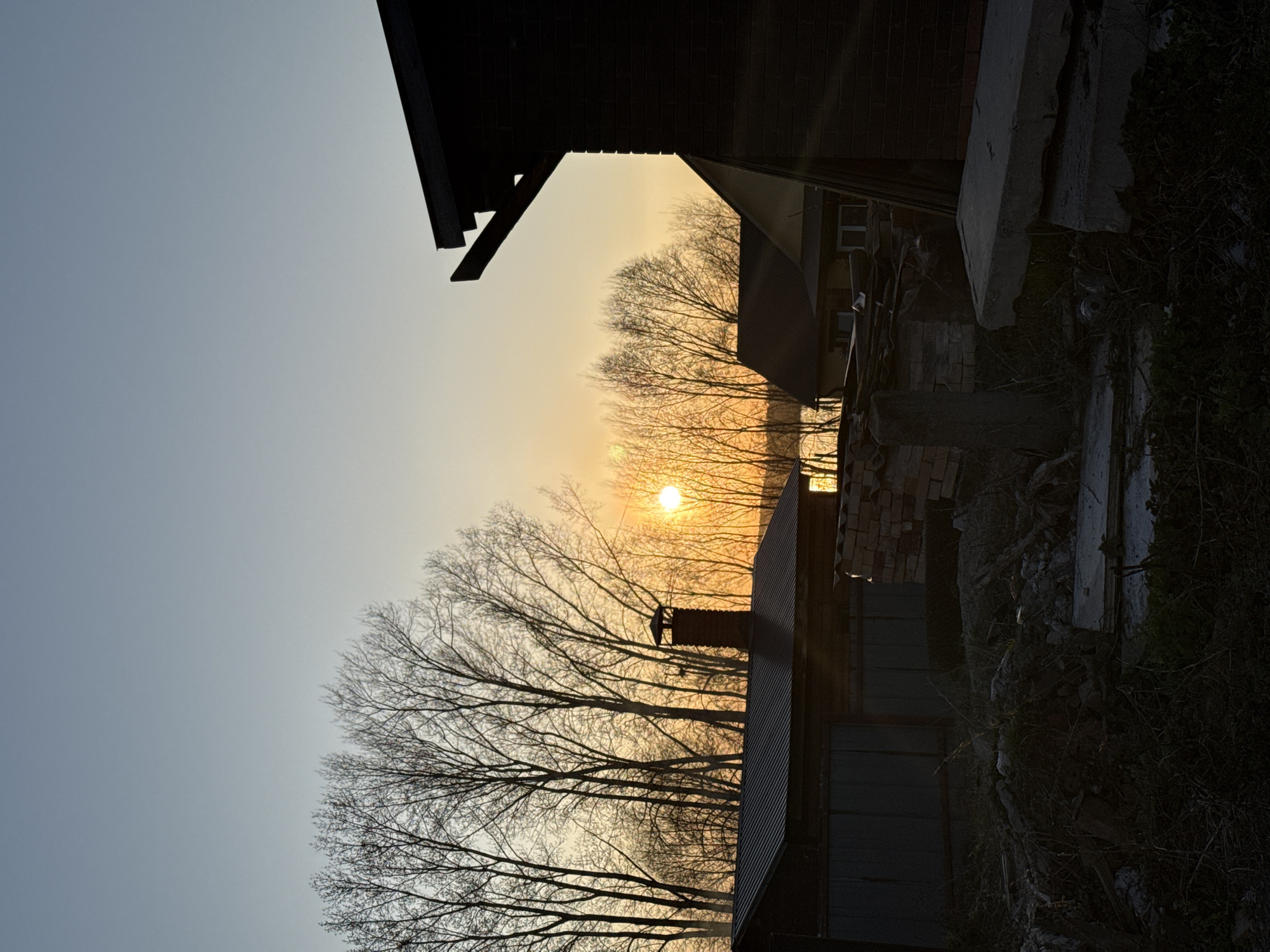 The image shows a landscape in what is potentially the city of Chaikovsky, Russia, taken at sunrise or sunset. Silhouetted buildings, including a brick structure and a shed with a chimney, frame a view of the sun setting/rising behind a line of bare trees. Debris and vegetation are visible on the right side of the image. The sky transitions from a pale blue to a golden hue near the horizon. There are no people visible in the frame.