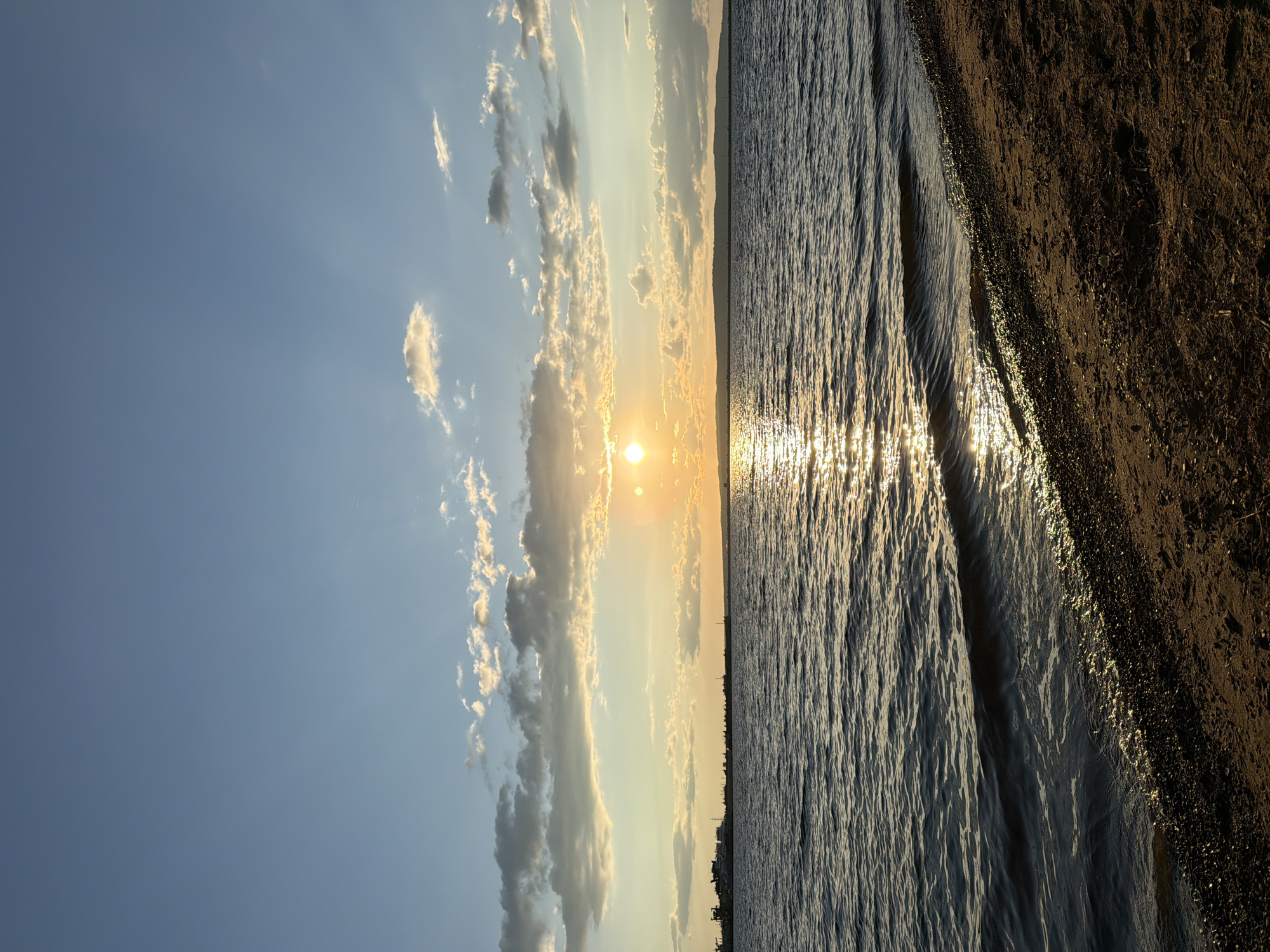 This image depicts a serene sunset over the Kama River in Chaikovsky, Russia. The sun, a bright orange orb, is partially obscured by scattered, wispy clouds, casting a shimmering golden path across the water. Gentle waves lap against a sandy and pebbly shoreline in the foreground. On the horizon, a line of dark hills is visible, and to the left, a collection of industrial buildings and docked ships suggest a port or industrial area. The sky is a gradient of pale blue to yellow and orange, with fluffy white clouds catching the last rays of sunlight. The overall scene evokes a sense of tranquility and the beauty of nature at dusk.