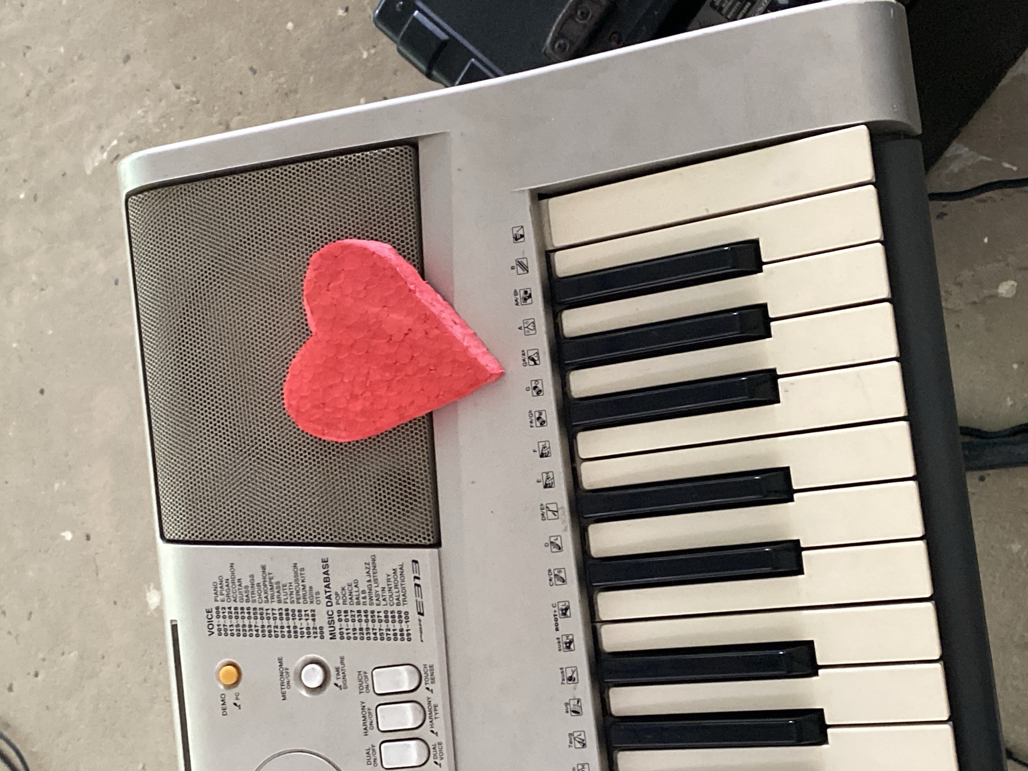 A top-down, close-up shot shows a portable electronic keyboard with a red, heart-shaped object placed on its left side. The keyboard is light gray with black and white keys. The keys are labeled with musical notes. The red heart appears to be made of styrofoam. The keyboard sits on what appears to be a gray surface. The keyboard model is a Yamaha E313.