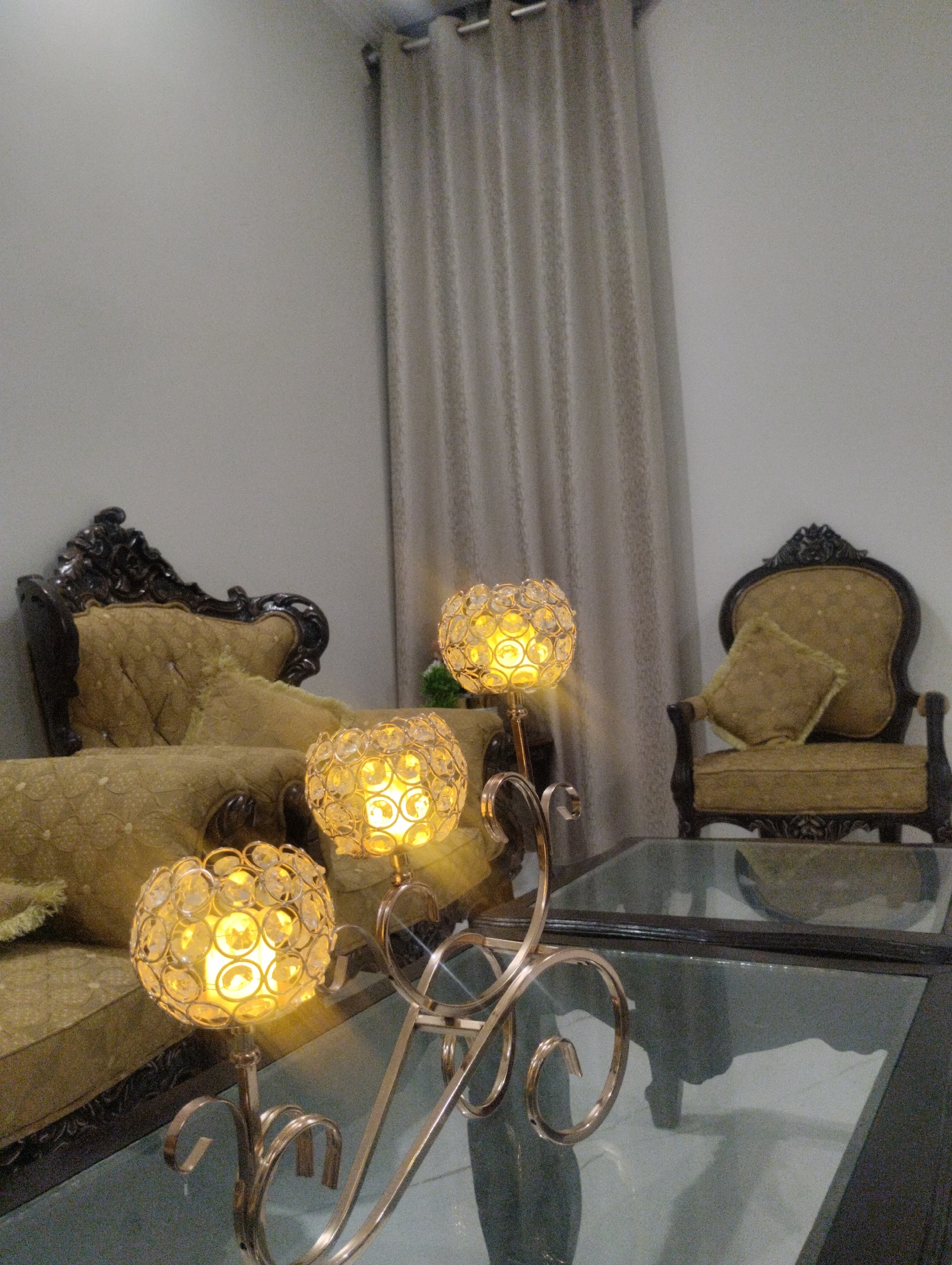 This media file depicts an indoor scene, likely a living room, with ornate furniture and decorative lighting. The subjects are primarily objects: a plush sofa with intricate detailing and a complementary armchair, both upholstered in a golden-yellow fabric with patterned accents. In the foreground, a modern glass-topped coffee table is present, and on it, a three-branched candelabra with spherical, crystal-like lamps. These lamps are illuminated, casting a warm, yellow glow and reflecting light. Behind the furniture, a pale, neutral-colored curtain hangs from a rod, suggesting a window. The overall aesthetic of the furniture is traditional and opulent, contrasting with the more contemporary design of the candelabra and coffee table. There are no people visible in the frame, and the scene appears to be staged for visual appeal rather than capturing a moment of activity. The lighting suggests it could be evening or the room is dimly lit, with the lamps providing the primary illumination. The background wall is a plain, light color, further emphasizing the focus on the furniture and decorative elements. No text is visible in the media file.