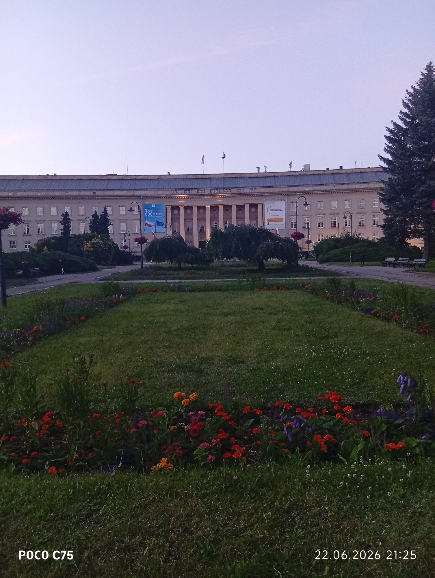 This media file depicts an exterior scene of a large, classical-style building, likely an institutional or governmental structure, with a prominent facade featuring columns and numerous windows. In front of the building, a park-like area is visible. This includes a well-maintained lawn with flower beds in the foreground, displaying a variety of colorful flowers, predominantly red and orange, with some purple accents. Several trees, including a large evergreen on the right and weeping trees closer to the building, add to the landscape. Benches are present along a paved walkway. Two flags are displayed on poles attached to the building, one appearing to be the Polish flag. A large banner is also visible hanging from the building, featuring text and graphics, though the details are indistinct. The time of day appears to be late evening or early dusk, given the soft, diffused light and the pale, clear sky. The weather seems calm and pleasant. The overall atmosphere is serene and peaceful, suggesting a public space. Visible text includes "POCO C75" and "22.06.2026 21:25" on the lower portion of the image, indicating the device used and the date and time the photo was taken. The location context is Wrocław, Poland.