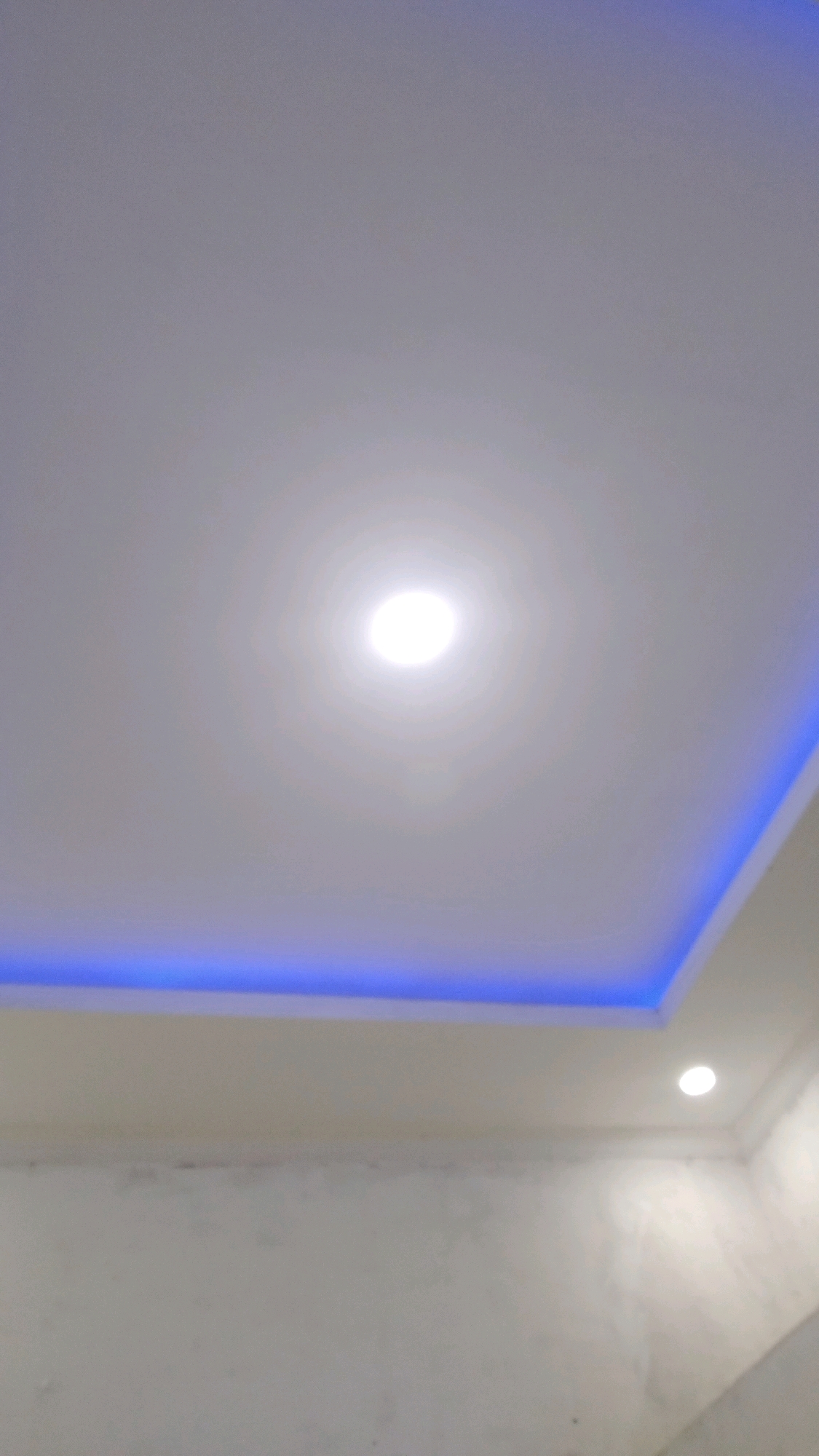 The image shows a modern ceiling with recessed lighting and cove lighting. A circular spotlight is in the center of the ceiling, and a band of blue LED lighting runs along the perimeter of a recessed section of the ceiling. The ceiling appears to be white and smooth. The lower portion of the image shows a portion of a wall, which is also white and appears to be textured. No people or specific objects are visible that would allow for a precise location within Kebon IX, Indonesia.