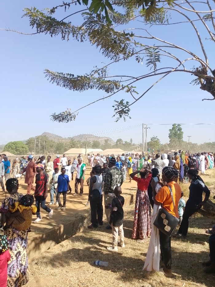 The image displays an outdoor gathering of approximately one hundred individuals, predominantly adults, on dry, light brown ground in the city of Kpak, Nigeria, under a clear, pale blue sky. In the foreground, a tree branch with green leaves extends from the upper left. A light blue plastic water bottle lies on the ground, mid-right. Several people are prominent in the foreground: a woman in a patterned dress carrying a child on her back, a young person in a black and red patterned shirt, a man in a white tank top, a child in a black long-sleeved shirt, a woman in a red top with a patterned skirt, and another woman in an orange t-shirt holding a white cloth. In the midground, a dense crowd of people, some wearing traditional attire, others in casual clothing, and at least one individual in a camouflage top, are standing and walking. Some members of the crowd hold green and white banners or flags. Low, single-story buildings are visible behind the crowd. The background features distant hills and mountains, interspersed with utility poles and power lines. No specific interactions beyond collective presence are discernible.