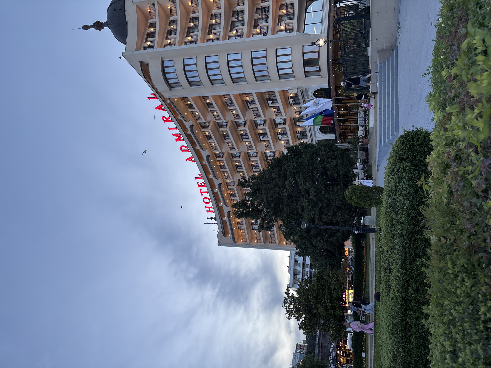 The image shows the Admiral Hotel in Varna, Bulgaria, as seen from a park. The hotel is a large, multi-story building with a curved facade and many balconies. Red neon lights spell out "HOTEL ADMIRAL" at the top of the building. There are several flags flying in front of the hotel, including the flag of Bulgaria. In the foreground, there is a green lawn with bushes and a pathway. A few people are walking in the park and on the steps leading up to the hotel. The sky is overcast, suggesting it is either early morning or late evening.
