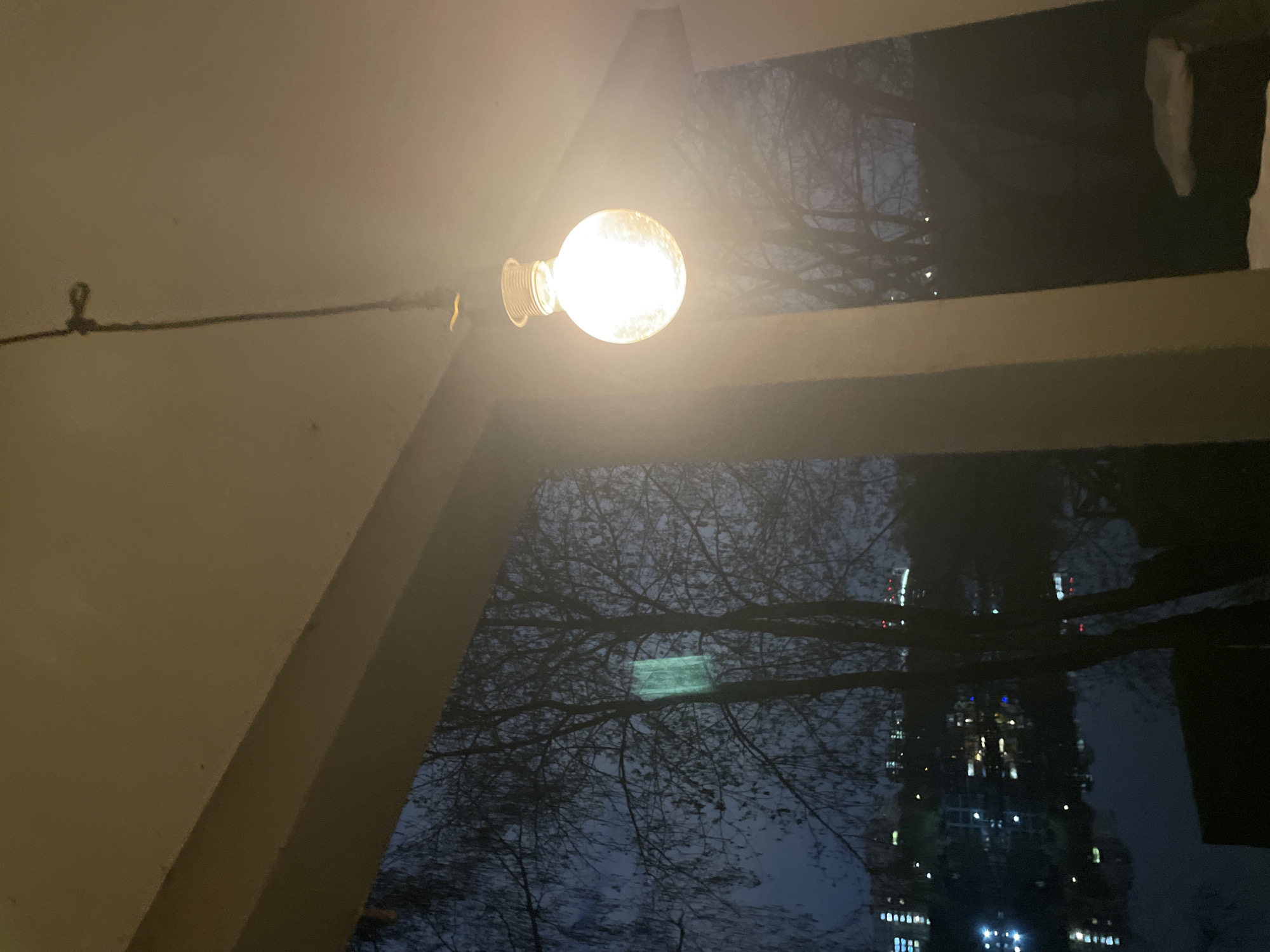 The image captures a nighttime view from what appears to be a balcony or an exterior part of a building, likely in an urban setting. A bright, spherical incandescent-style light bulb hangs prominently in the upper center, casting a warm, yellowish glow. It is suspended by a dark cable or wire, which extends to the upper left and is secured to a light-colored wall or ceiling by a small metal hook.
The foreground is defined by beige or off-white architectural elements, possibly beams or parts of a building's structure, which frame the scene with strong diagonal and horizontal lines. Beyond these structures, the bare, dark branches of trees are silhouetted against a cool, dark blue-grey night sky, suggesting either autumn or winter.
Below the light bulb and the immediate architectural framework, a reflective surface—perhaps a large window, calm body of water, or a very glossy surface—mirrors the surrounding environment. This reflection shows more tree branches and, notably, a distant, tall urban building. This high-rise is illuminated with numerous lights, appearing as glowing white, yellow, red, and blue rectangles and streaks against its dark exterior. A faint, rectangular, greenish light is also visible reflected among the tree branches.
No people or specific activities are depicted, contributing to a serene and still atmosphere. The dominant light sources are the foreground bulb and the distant city lights. The scene evokes a sense of urban solitude at night, with the natural elements (trees) coexisting with man-made structures. No visible text is present in the image. The location context for this scene is Moscow, Russia.