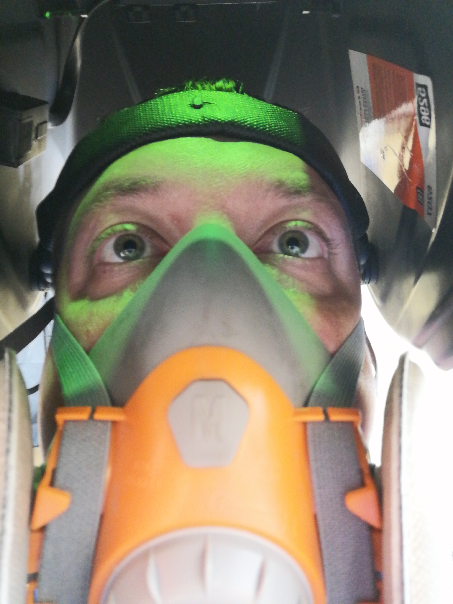 A close-up, upward-facing perspective shows a person wearing a full-face respirator. The respirator is primarily grey and orange, with green straps. The person's face, illuminated by a green light, is visible above the mask. Their eyes are open and appear to be looking directly at the viewer. Above the person, dark objects form the background. A sticker with text "OSSA" is visible on the right side of the upper background. The location is Unknown, Unknown.