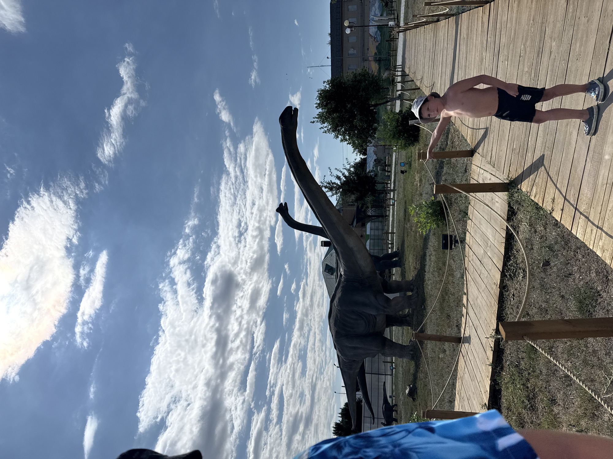 The image depicts an outdoor scene at the Sol-Iletsk Dinosaur Park in Sol-Iletsk, Russia. In the foreground, a young boy, shirtless and wearing a hat and shorts, stands on a wooden boardwalk and points to his left. He is wearing dark shorts with a white stripe and grey shoes. The boardwalk extends into the background, lined with wooden posts and ropes. To the left of the boardwalk, several large dinosaur statues are visible, including two brachiosaurus figures with long necks. The ground is covered in dry grass, and there are some trees and bushes. In the background, buildings and a fence can be seen. The sky is blue with scattered white clouds, and the sun is shining. The overall impression is of a sunny day at a tourist attraction.