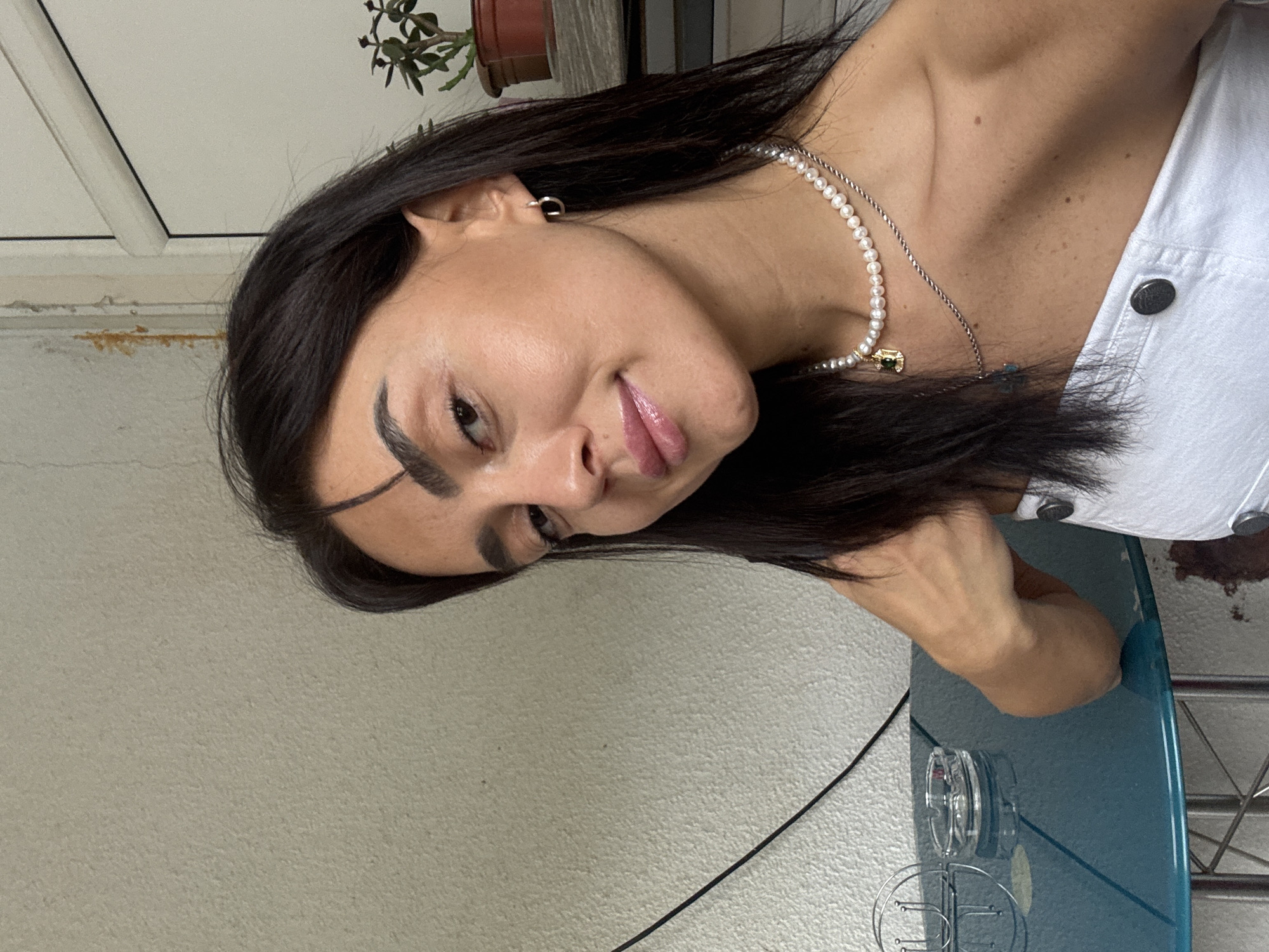 A young woman with dark hair and dark eyes is captured in a selfie taken outdoors in Tbilisi, Georgia. She is leaning forward, her head tilted slightly to her left, with a soft smile on her face. She is wearing a white strapless top and two necklaces, one a string of pearls with a gold pendant and the other a thinner silver chain. She also has small hoop earrings. The background shows a textured, off-white wall with some visible peeling paint and staining near the top. To the right, a white doorframe and a potted plant with green leaves are visible. The woman is seated at a round, teal-colored table. On the table are an ashtray and a metal object that appears to be a napkin holder. The overall setting suggests a casual outdoor seating area.