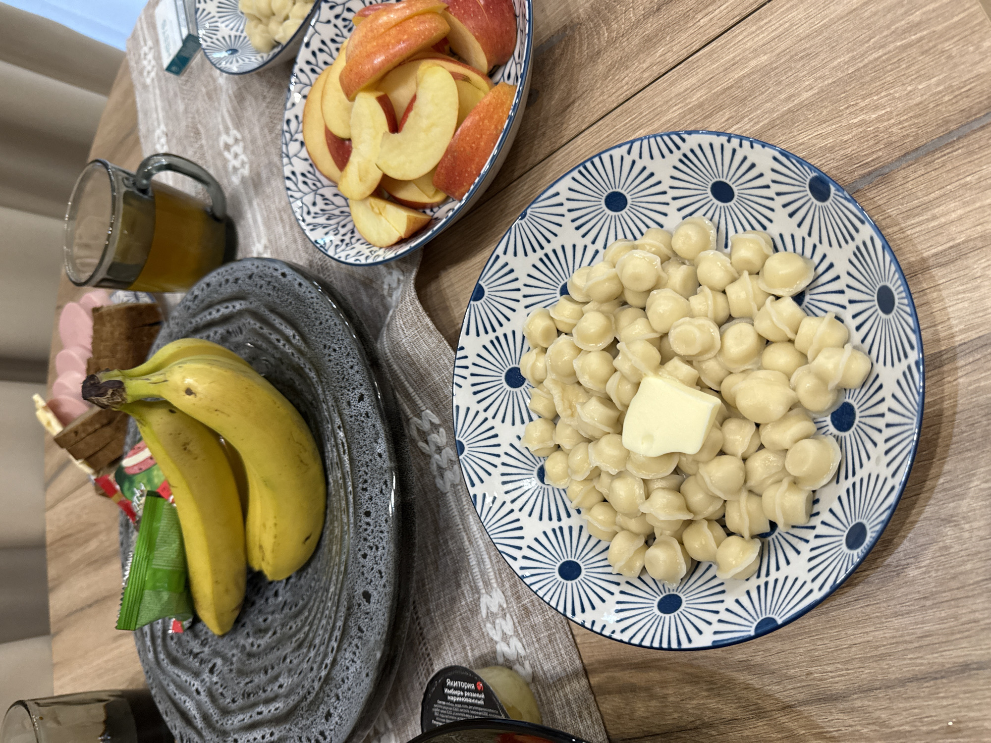 The image depicts a breakfast scene on a wooden table. The location is likely Buzhaninovo, Russia, based on the Cyrillic text on a container. A plate of dumplings topped with butter sits in the upper right, alongside a bowl of sliced apples. To the left, a plate holds bananas and a glass contains juice. The tablecloth is patterned.