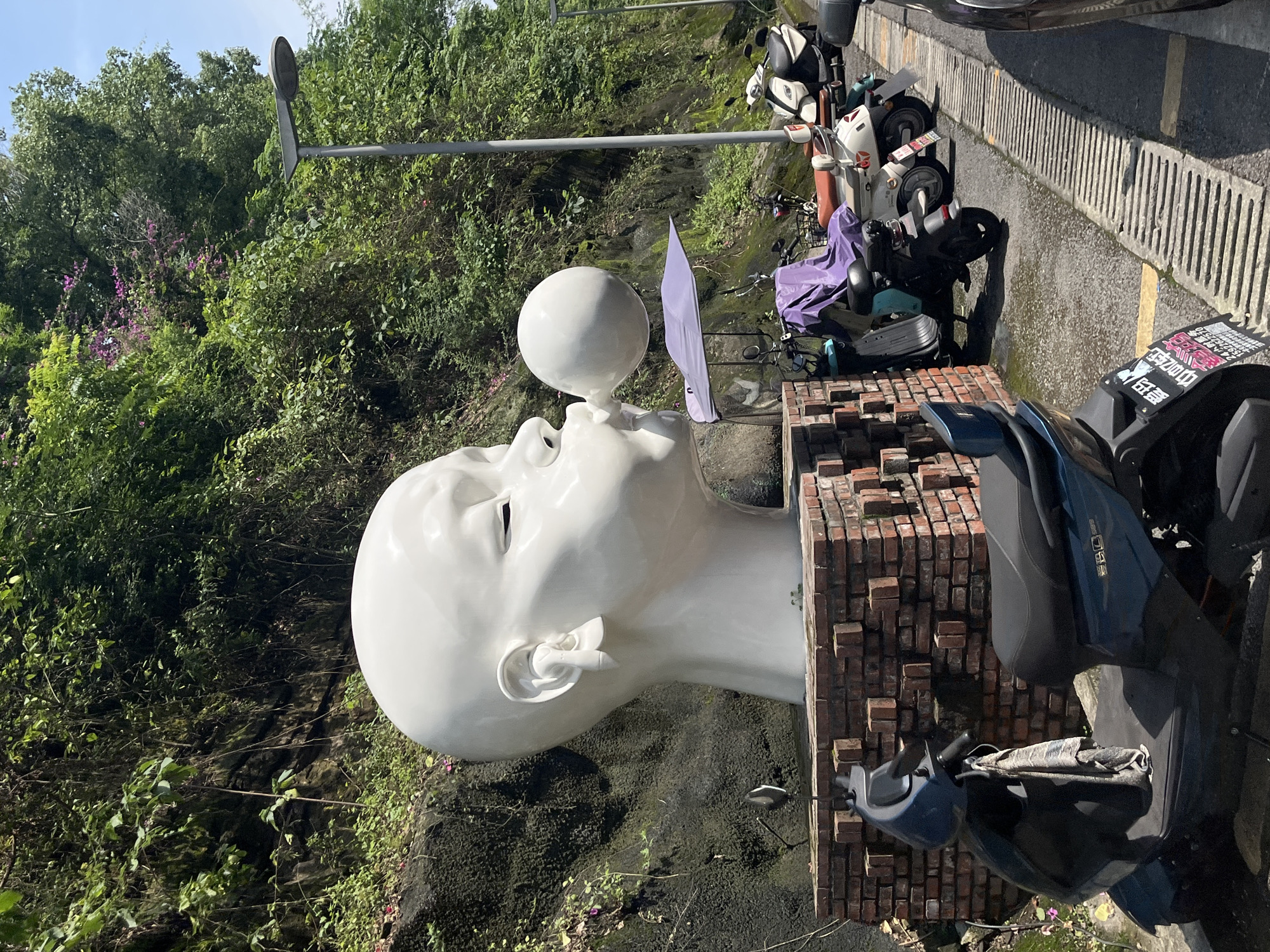 The image shows a large white sculpture of a person's head with a bubble gum bubble being blown out of its mouth, located in a city park. The sculpture is situated in front of a rocky, overgrown hillside. In the foreground, several scooters are parked along a paved path next to a drain grate. A lamppost stands to the right of the sculpture. The environment suggests an outdoor public space, possibly a park or roadside area. The style of the sculpture and the presence of the scooters indicate an urban setting in China. Based on the provided context, this scene is located in the Chongqing High-tech Zone, China.
