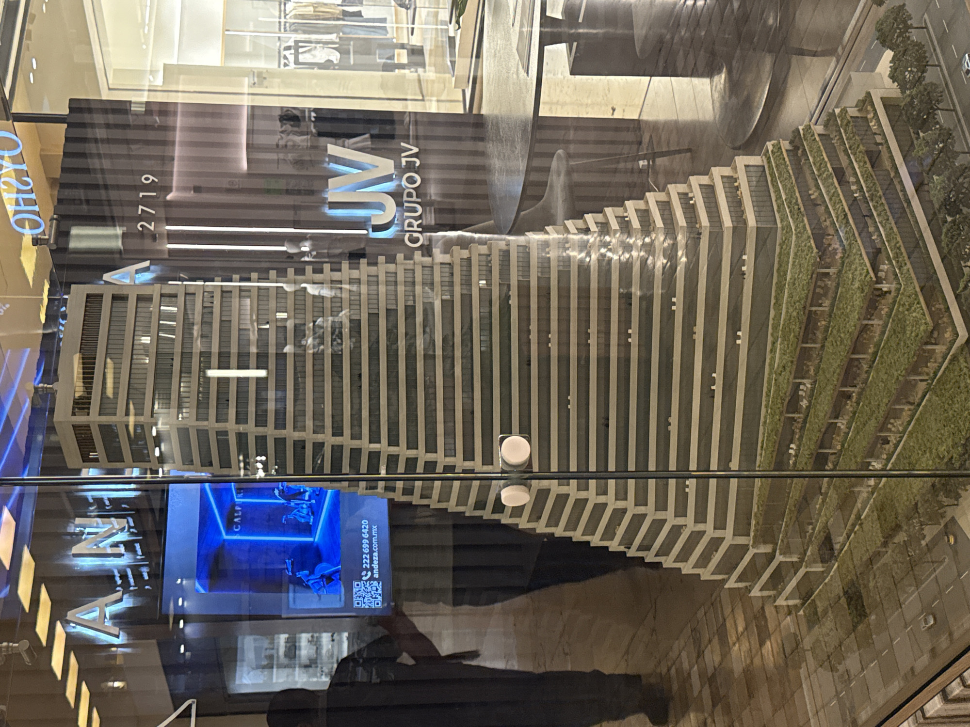 The image captures a detailed architectural model displayed in a storefront window. The model represents a high-rise building characterized by terraced levels that incorporate green spaces. The building’s design includes vertical lines and tiered sections with greenery visible on some levels.
Reflections in the window overlay the model, including parts of the storefront signage, the building number "2719," and the words "GRUPO JV." A screen with a fitness class advertisement is also visible in the reflection.
Beneath the model and its reflections, part of the observer is captured in the reflection, likely a person taking the picture.
The location is suggested to be in the city of Puebla, Mexico.