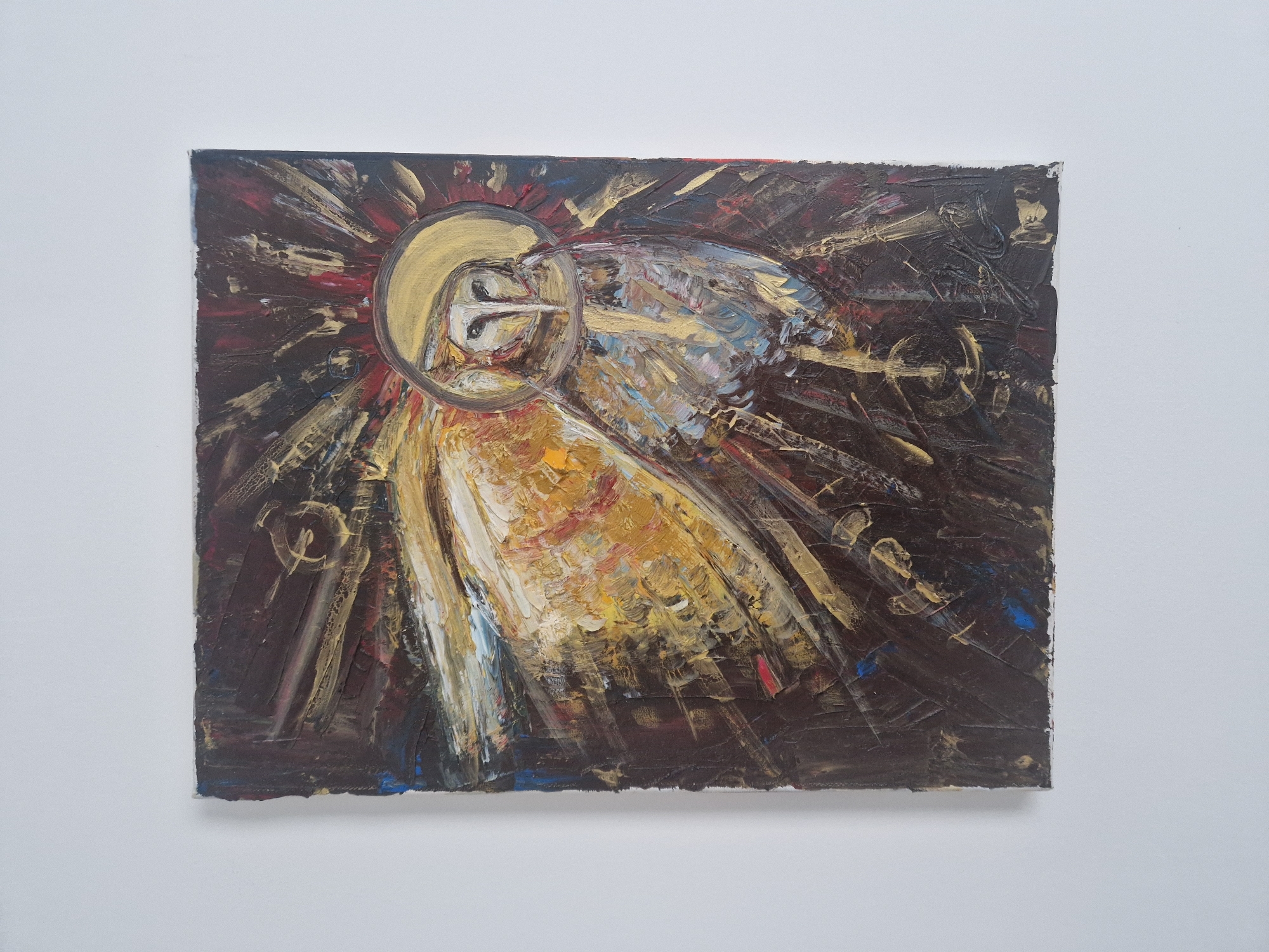 A vertical rectangular painting is centrally mounted on a plain white wall in Unknown, Unknown. The painting, rendered with a heavy impasto technique, depicts a stylized barn owl with a golden halo around its head. The owl's face is pale and pointed, while its body features textured brushstrokes in shades of white, gold, blue, and brown. Radiating outwards from behind the owl is a burst of light, characterized by thick, expressive brushstrokes in gold, yellow, and red. This central motif is set against a dark, textured background primarily composed of deep browns and blacks, with hints of red and blue. The edges of the canvas appear raw and slightly frayed. The wall provides a stark, uncluttered backdrop for the artwork.