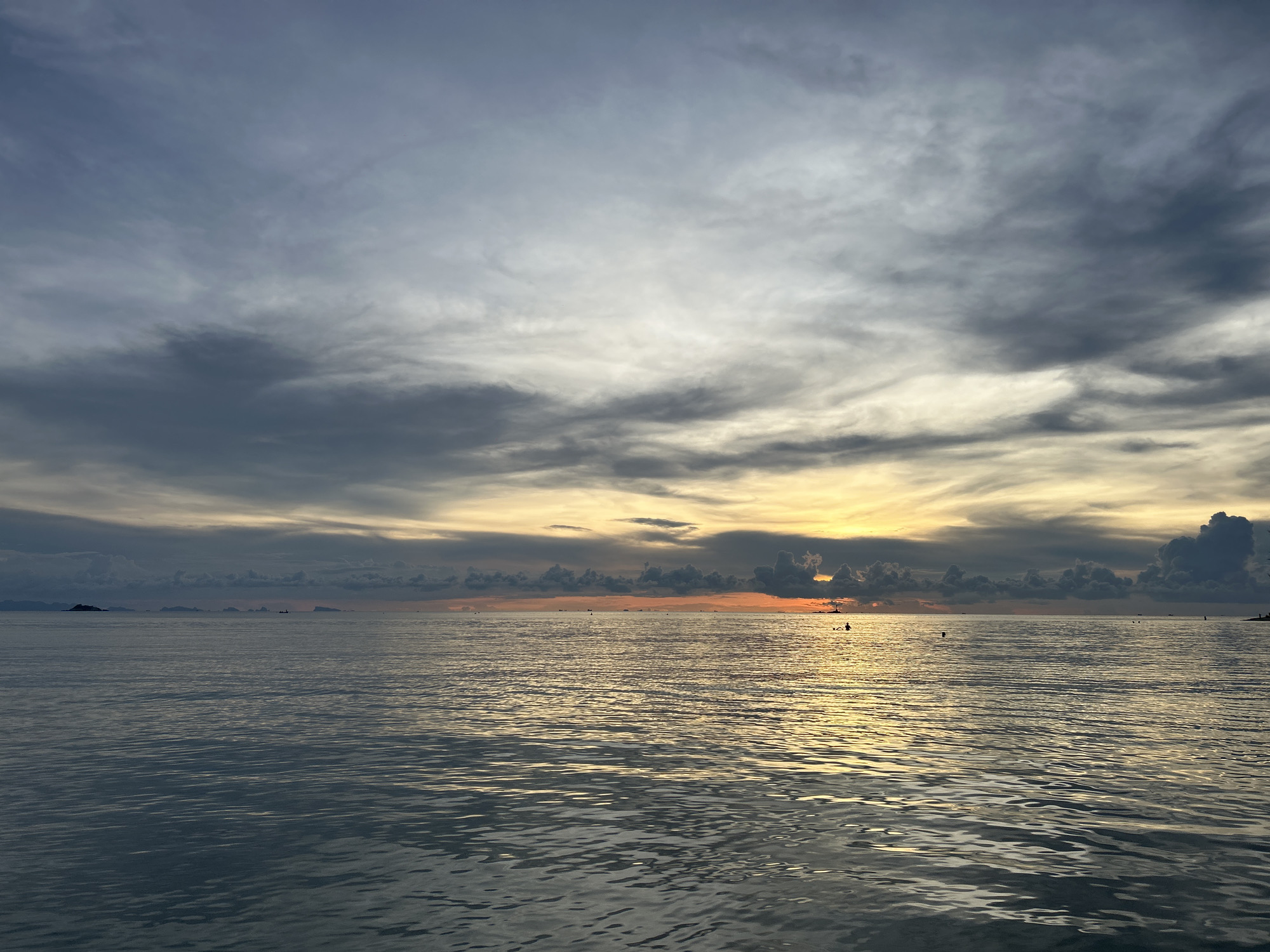 The image depicts a seascape at sunset, likely near Baan Sri Thanu, Thailand. The sky is filled with striated clouds, transitioning from dark gray above to a band of orange and yellow near the horizon. The sea reflects the colors of the sky, with golden sunlight shimmering on the water's surface. In the distance, several small islands and boats are visible. The overall atmosphere is tranquil and subdued.