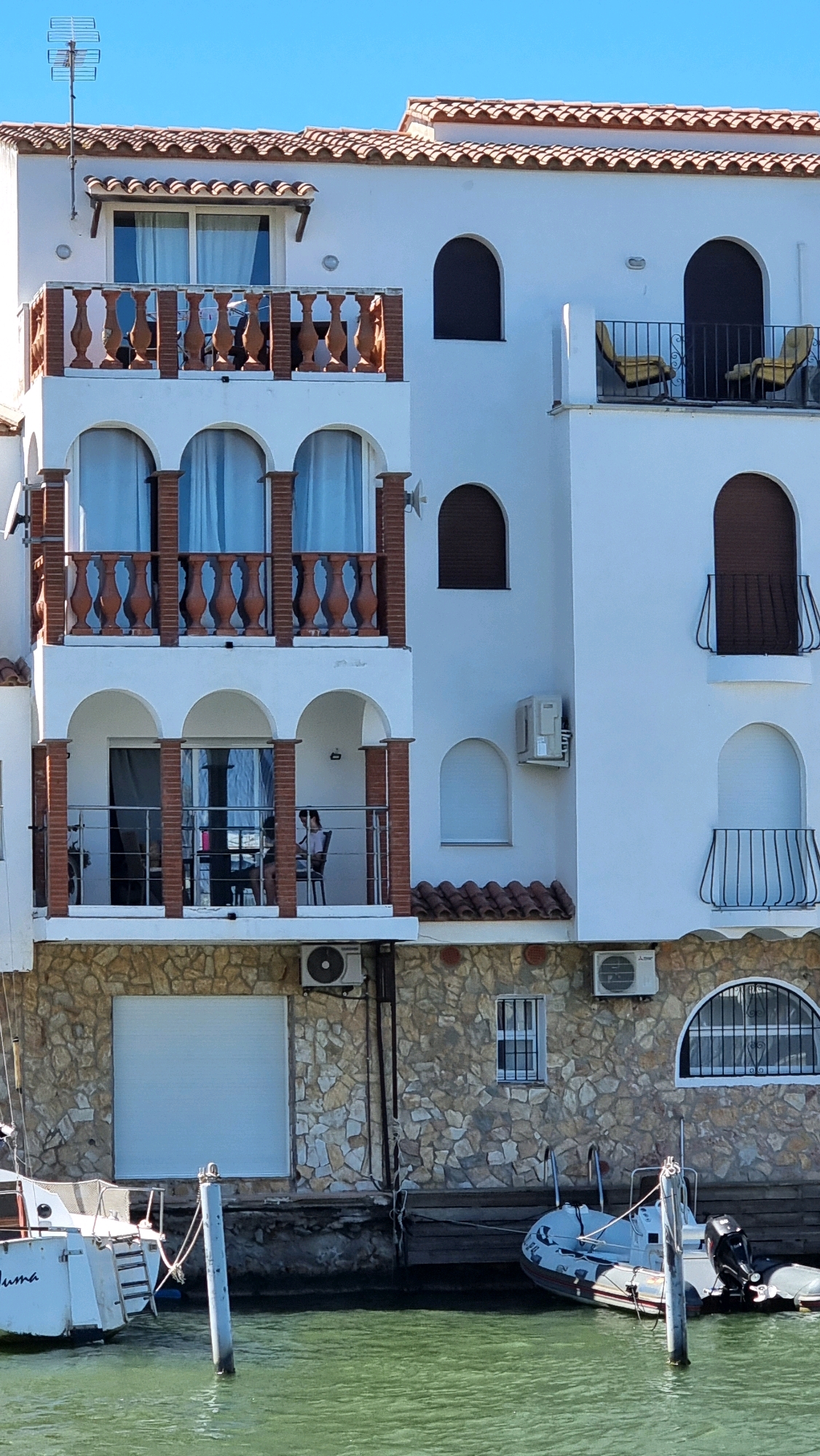 The image shows a white multi-story building with a red tiled roof, situated next to a body of water, likely a canal or harbor in Castelló d'Empúries, Spain. The building features several balconies with decorative terracotta balustrades and metal railings. Arched windows and doorways are visible, some with dark shutters. Two boats are docked in the water in front of the building. One boat on the left has "LUMA" written on its stern. An air conditioning unit is mounted on the exterior wall of the building. A person is seated on a balcony on the second floor. The environment appears to be a sunny day with a clear blue sky.