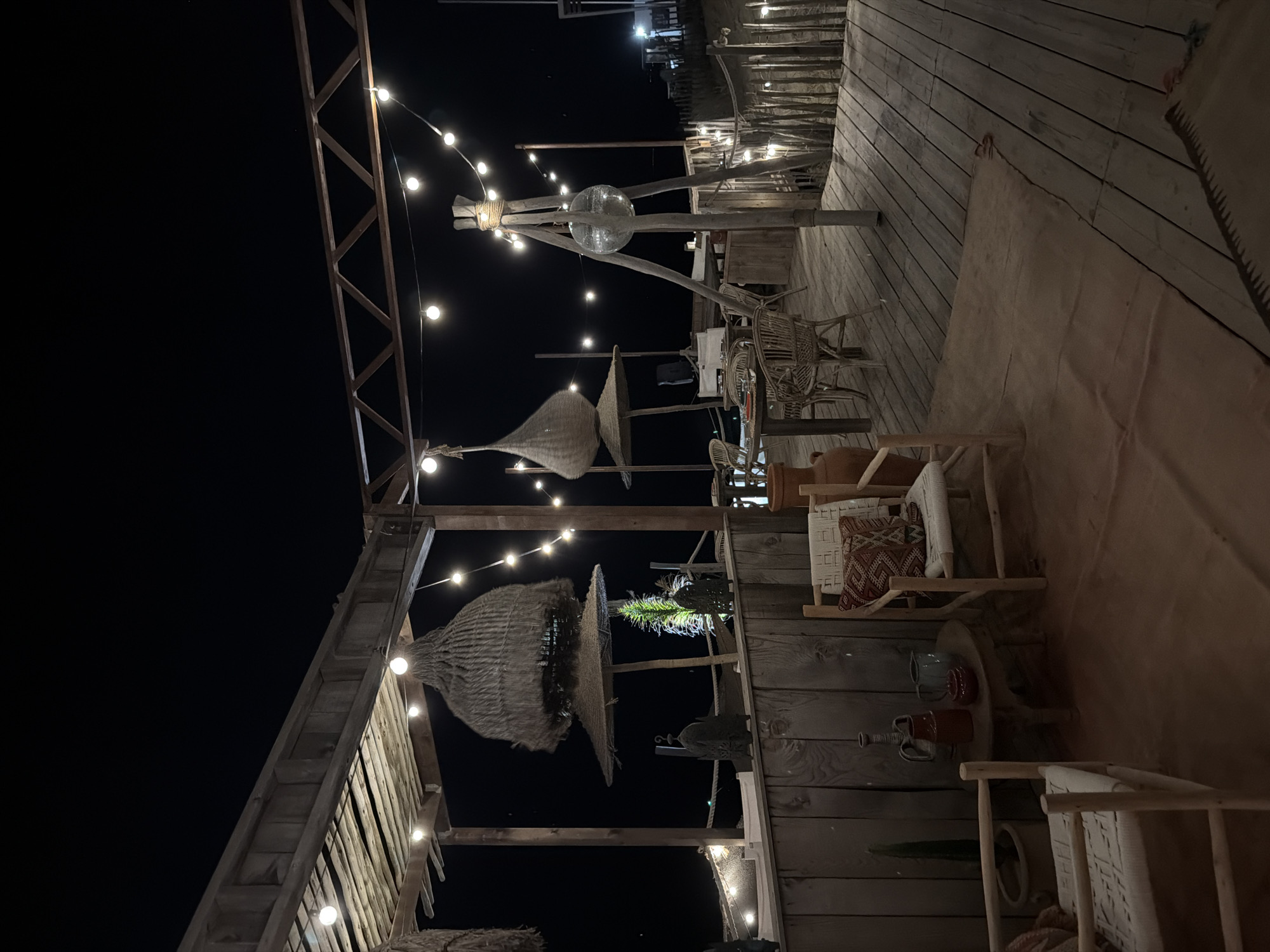 This is a low-angle, full-frame, vertical, outdoor night shot of a rustic, open-air restaurant or bar area in Torreilles, France. The scene is illuminated by string lights and appears to be situated near a beach or water. The foreground features a wooden deck with a textured, rust-colored rug. Two simple wooden chairs with woven seats and patterned cushions are visible. To the left, a small round wooden table holds decorative pottery and a potted plant. Further back, several more wicker chairs and tables are arranged under a thatched-roof canopy. In the background, a disco ball hangs from a wooden tripod, surrounded by more string lights. The overall aesthetic is bohemian and relaxed, with natural materials like wood and straw dominating the scene. The dark sky suggests it is nighttime. There are no people visible in the image.