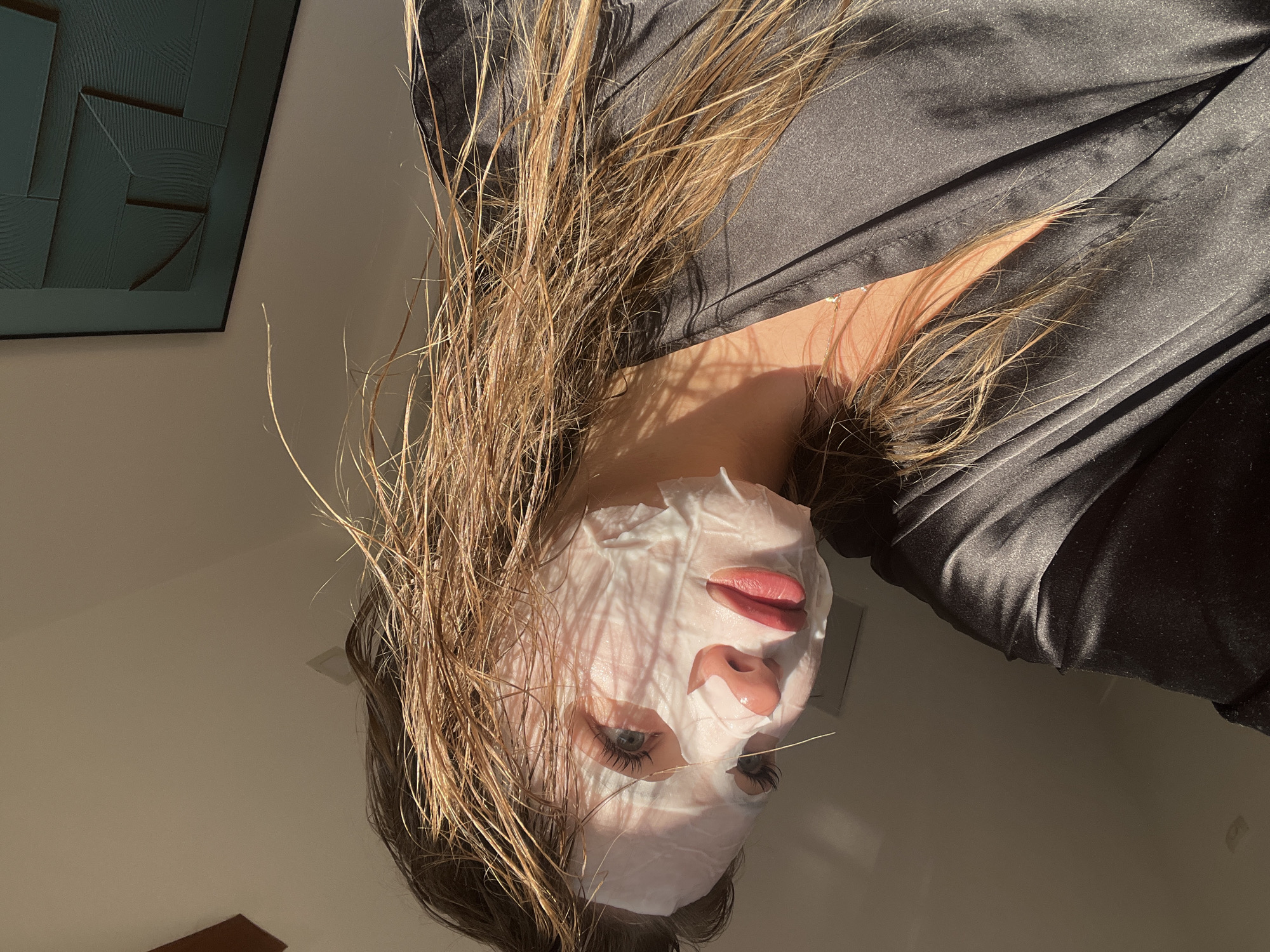 A low-angle, slightly rotated, close-up indoor shot shows a woman wearing a white sheet facial mask, looking at the camera. Her light brown hair is loose and wet, partially obscuring her face. Only her eyes, lips, and nose are visible through the mask. She is wearing a dark-colored, silky robe. A framed artwork with geometric designs hangs on the wall in the background. The location is uncertain. Sotonići, Montenegro is not identifiable in the scene.