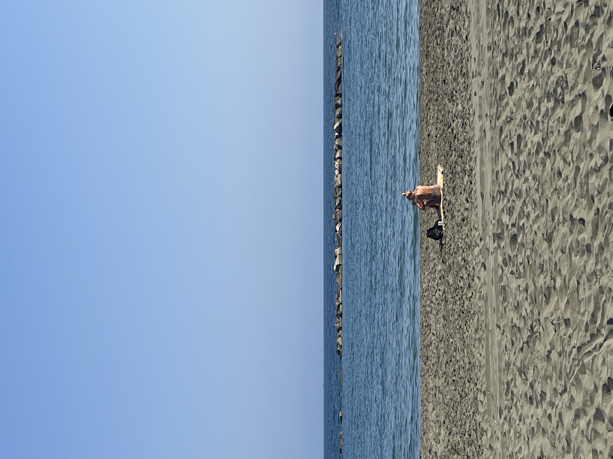 A person is sitting on a sandy beach in Agios Tychona Community, Cyprus. The person is facing away from the camera, with their back to the viewer, and appears to be wearing a swimsuit. They are seated on a light-colored towel or mat. Next to the person, on the sand, is a black bag or backpack. The beach is composed of coarse sand with visible footprints and ripples. The sea is calm and blue, stretching to the horizon where it meets a clear, pale blue sky. A breakwater, constructed of large rocks, extends horizontally across the water in the mid-ground of the image. The overall scene is one of tranquility and a summer day at the beach.