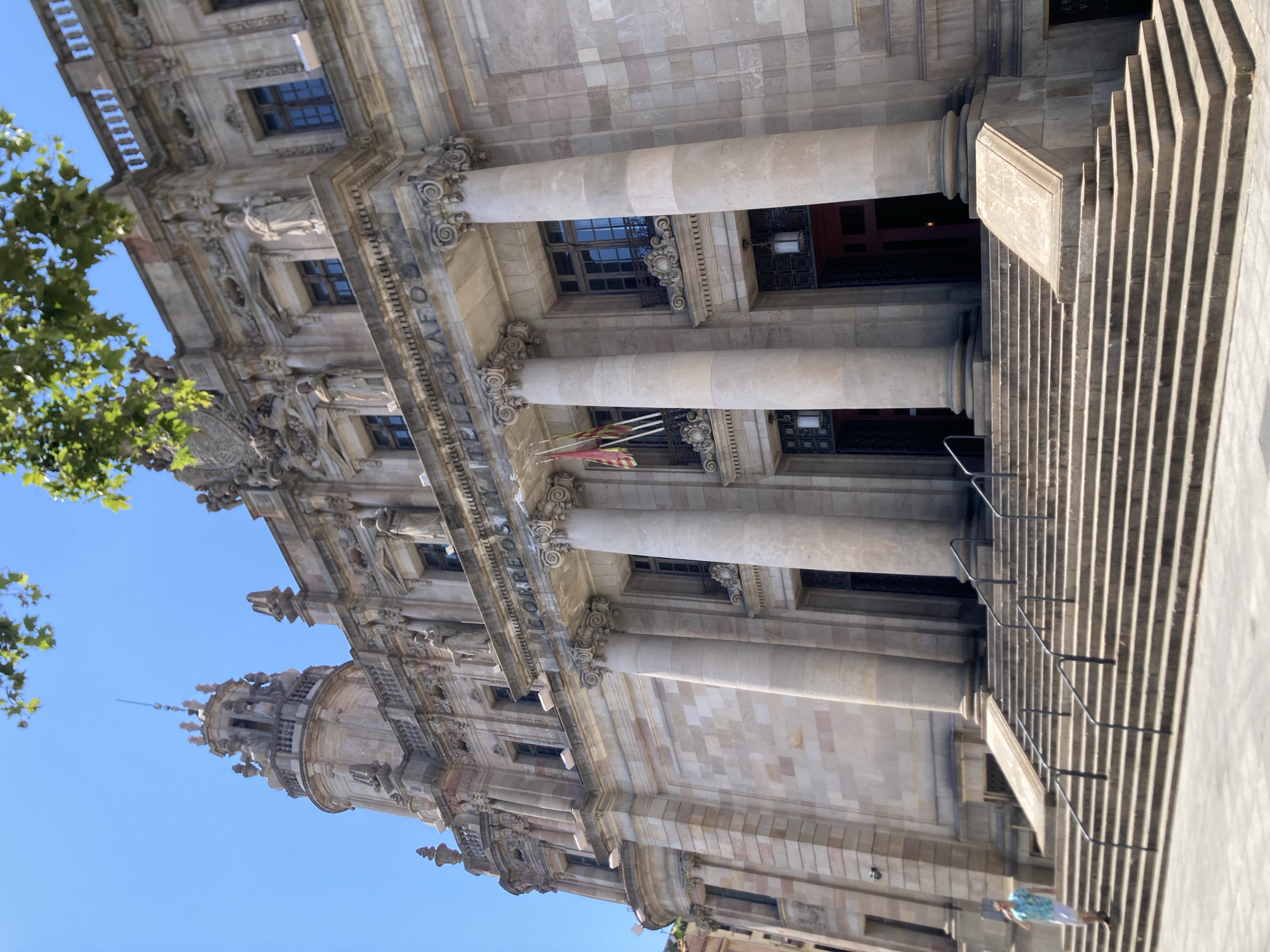 The image shows a low-angle, sideways view of an ornate stone building, likely a historic public building in Barcelona, Spain. The architecture features classical elements such as large columns with decorative capitals, intricate carvings, and multiple arched windows. A grand stone staircase with handrails leads up to the entrance on the right side of the frame. Green foliage from a tree is visible on the left. In the distance, a tower with spires can be seen.
A person wearing a patterned shirt and white pants is standing at the bottom of the stairs, looking towards the building. Another person is partially visible on the stairs. The scene appears to be outdoors during daylight, under a clear blue sky. The sunlight casts distinct shadows from the stairs and the architectural details of the building, suggesting it is midday or afternoon.
Visible text on the facade reads "TELEGRAFOS", identifying the building as the former main telegraph office in Barcelona. The overall impression is one of grandeur and historical significance, with a sense of an ordinary day unfolding in front of a monumental structure.