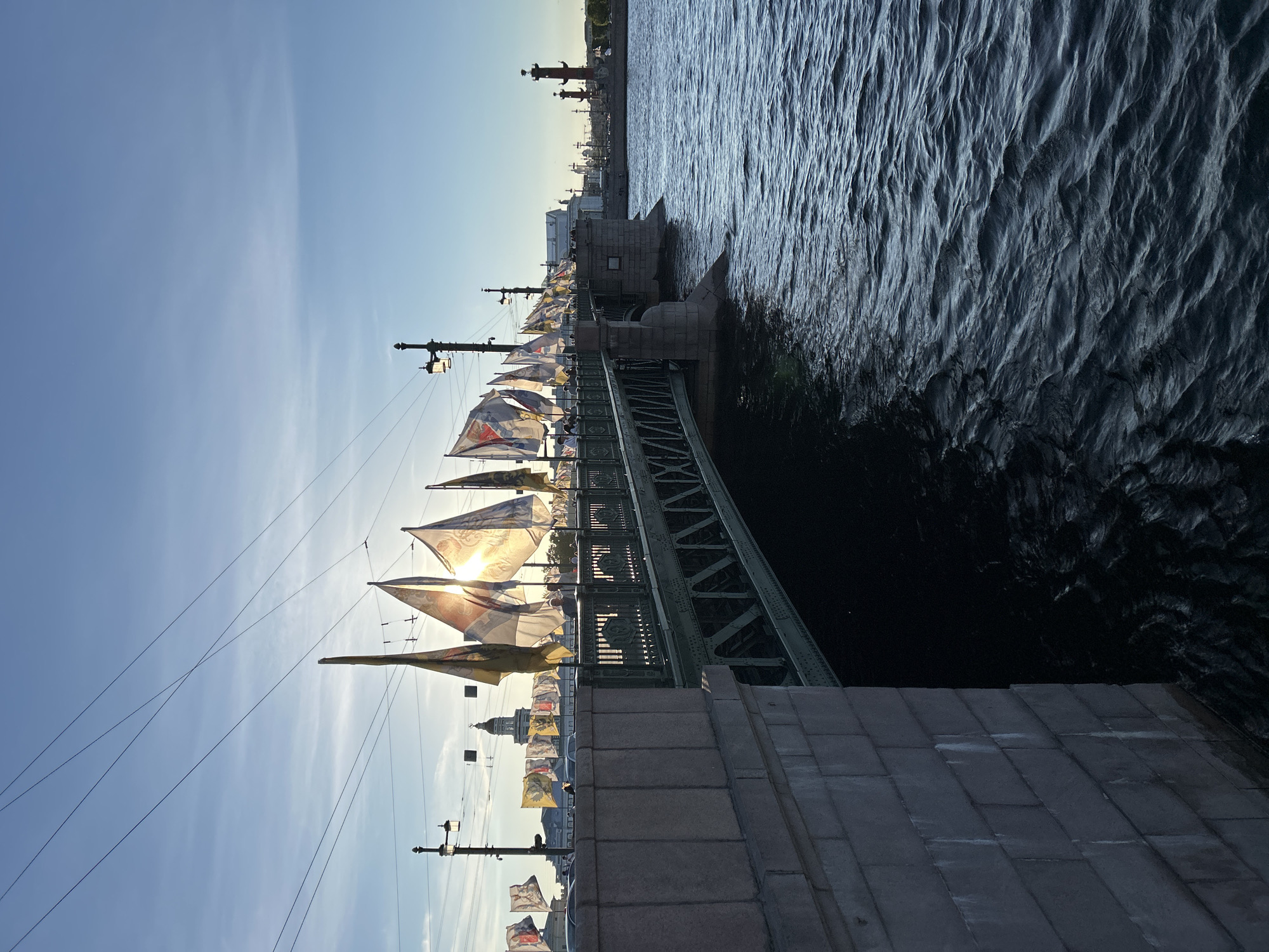 In a city scene under a clear blue sky with wispy clouds, a green metal bridge spans a dark, rippling body of water. The bridge, supported by stone abutments, is adorned with numerous flags that flutter in the breeze. Several streetlights with intricate tops are visible along the bridge's railing. In the background, a tall domed building with a spire and a tall red column with a sculpture on top are prominent landmarks. The sun is low in the sky, casting a warm glow and creating lens flare as it shines through some of the flags. A few cars are visible on the bridge, and a couple of individuals are standing near the railing. The water reflects the sky and the bridge structure, with noticeable waves disturbing the surface. The overall atmosphere suggests a late afternoon or early evening setting in a historically significant urban environment.