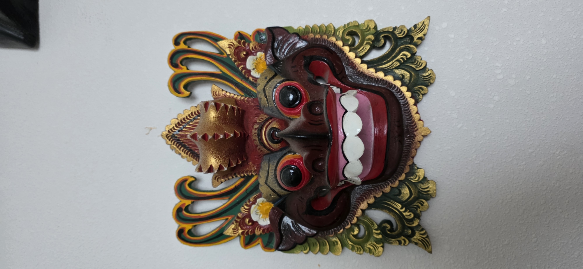 A carved and painted wooden mask is centrally displayed against a textured, off-white wall. The mask depicts a creature with a dark brown face, large black pupils, red and orange irises, and a wide, smiling mouth revealing white teeth and pink gums. A prominent dark brown nose is situated between the eyes. Above the face, a multi-layered, crown-like headpiece is rendered in gold and red, featuring textured triangular patterns. Elaborate, stylized scrollwork and tendrils, painted in gold, dark green, and yellow, extend outwards from the sides and top of the mask, framing the central face. Small white and yellow floral elements are integrated into the decorative scrollwork near the forehead. The entire mask exhibits vibrant, glossy paint finishes. The wall behind the mask is uniformly colored and shows a fine, speckled texture. A small, faint, L-shaped mark is visible on the wall above the upper right portion of the mask.