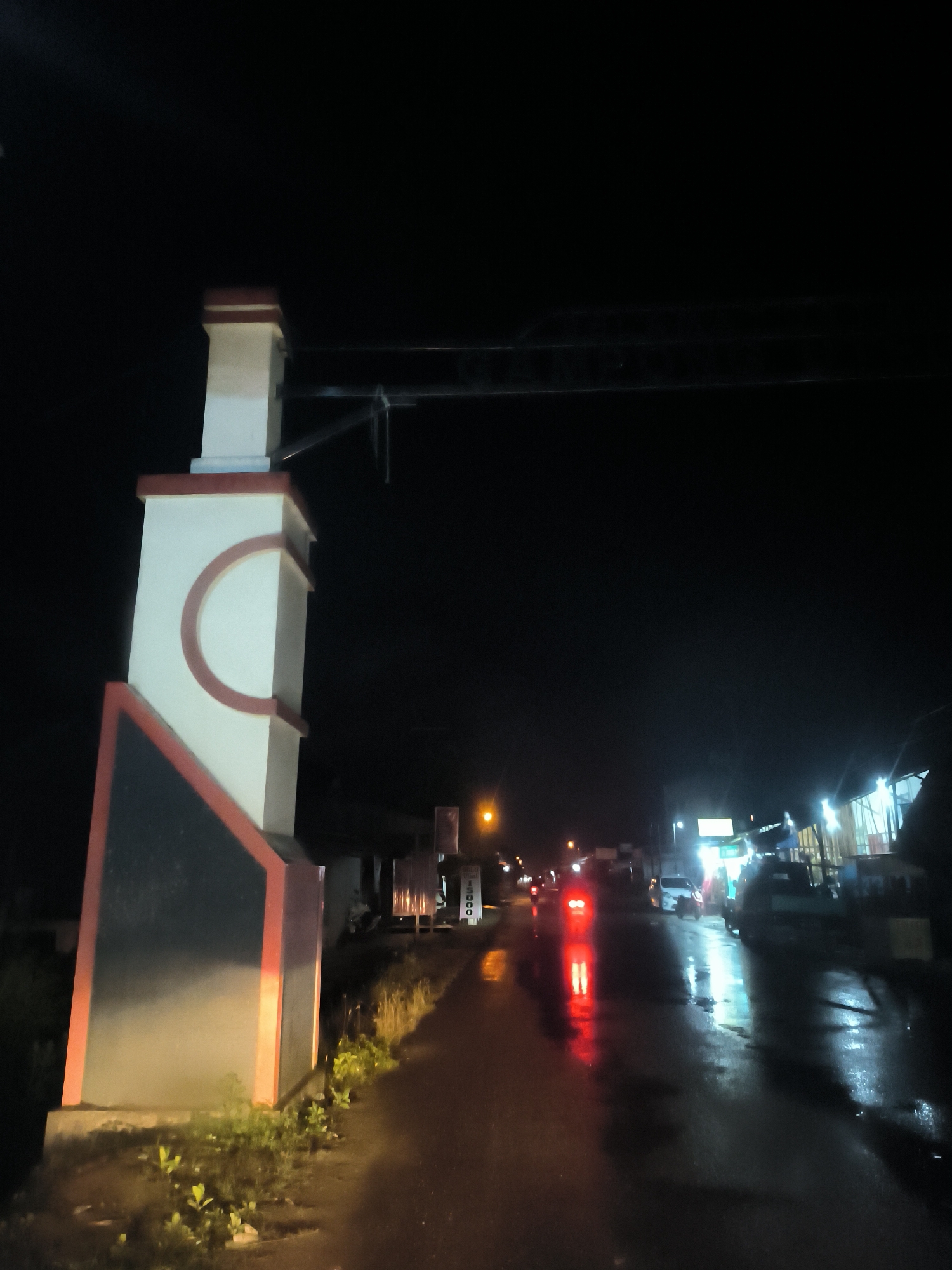 The image depicts a nighttime scene on a wet road in Lhokseumawe, Indonesia, suggesting recent rain. In the immediate foreground on the left, a tall, decorative pillar stands prominently. It is white with distinctive reddish-orange accents, including a large C-shaped design on its side, and a reddish base and top. Small patches of green vegetation are visible at the pillar's base.
The road, stretching into the distance, is reflective due to its wet surface. Bright red reflections from the taillights of a vehicle are visible near the center of the frame, indicating traffic. Further down, a white vehicle and other indistinct shapes suggest more cars. Along the right side of the road, several simple buildings or stalls are illuminated by bright white and yellow artificial lights, which also reflect on the wet pavement.
The sky above is dark, contrasting with the light sources below. On a signpost located on the right side of the road, near the foreground pillar, the numbers "5000" are clearly visible, possibly indicating a price. Below "5000", the text "IBU" or "BU" can be discerned. The scene conveys a quiet, illuminated street atmosphere at night.
