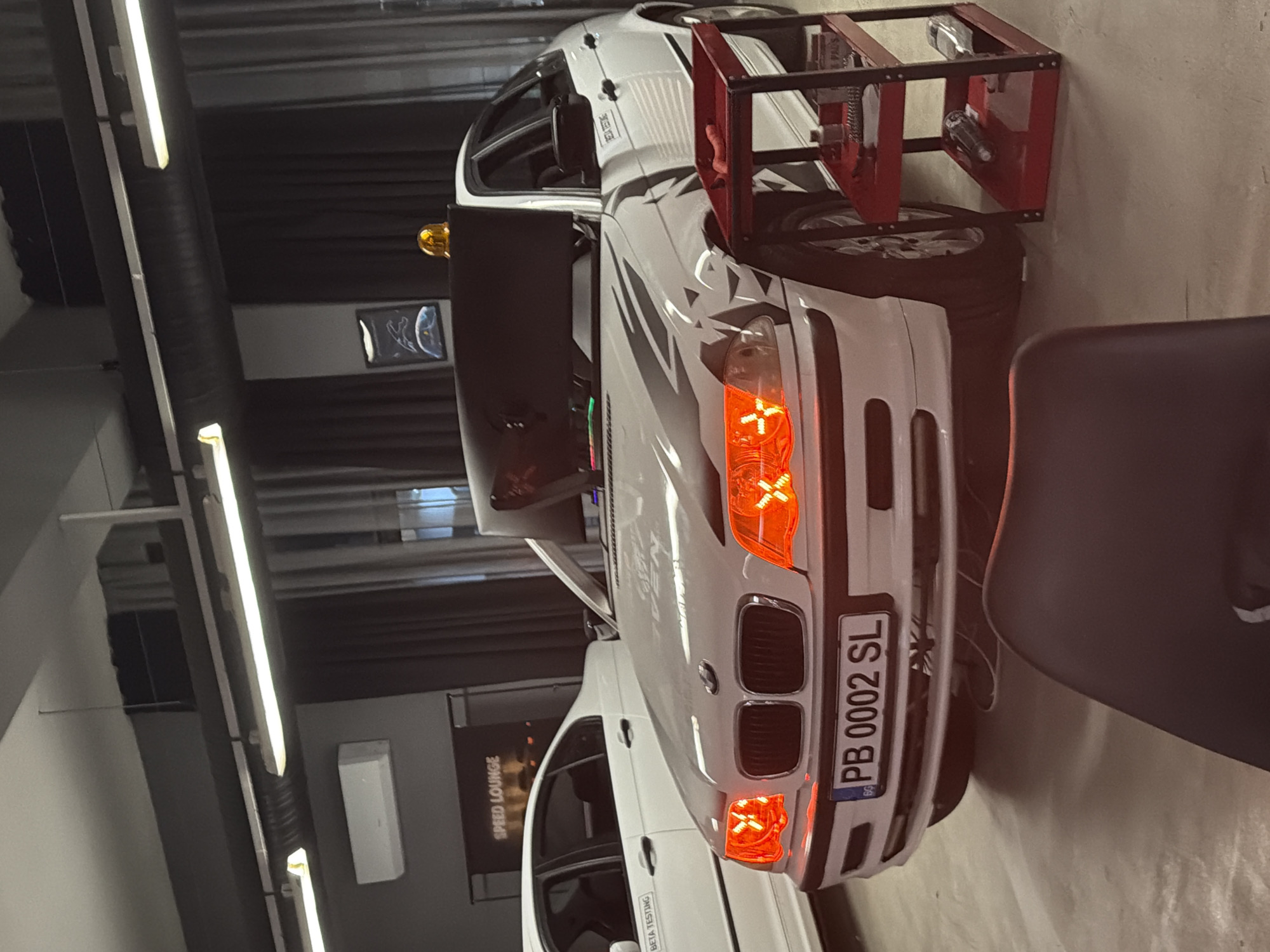 The image shows the front of a white BMW E46 series car with Bulgarian license plate "PB 0002 SL" parked inside a facility that seems to be related to car testing or modifications. The location is suggested to be in Plovdiv, Bulgaria, as the license plates with "PB" abbreviation are common for the city. The car is surrounded by equipment, including a red metal rack. The scene appears to be indoors, possibly in a speed lounge or car workshop, with lighting from fluorescent lamps. The environment suggests a professional automotive testing or customization setting. A dark chair is also visible on the right side of the frame.