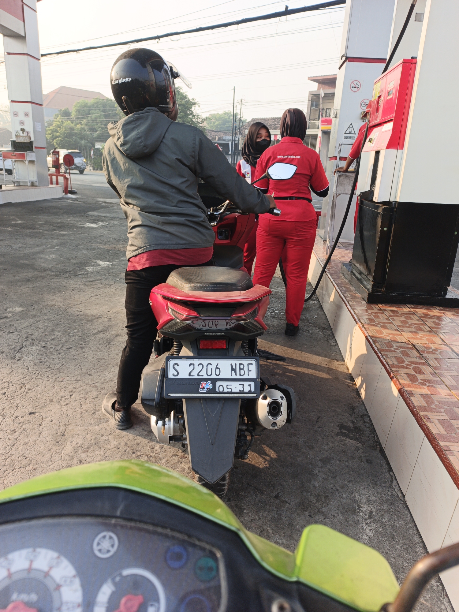 In Pacing, Indonesia, a person wearing a black helmet and a gray hooded jacket sits on a red motorcycle at a gas station. They are positioned in front of two female gas station attendants, one in a black hijab and black mask, and the other in a red uniform. The attendant in the red uniform is tending to a gas pump. The motorcycle's license plate is visible, reading "S 2206 NBF". The ground is paved with asphalt, and the gas station has tiled flooring along the pump area. The background shows a street with a white car and buildings. The scene is illuminated by natural daylight, with the sun casting shadows.