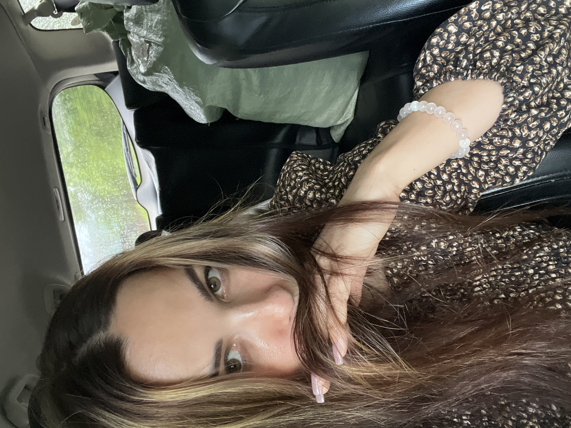 A young woman with long, brown and blonde-highlighted hair sits in the back seat of a car, holding a portion of her hair in front of her face. She is wearing a dark brown dress with a light brown and white speckled pattern. On her left wrist, she wears a bracelet made of light pink and white beads. A large, light green duffel bag is visible in the seat behind her. The background shows the rear window of the car, with a blurred view of green foliage and what appears to be rain or water droplets on the glass.