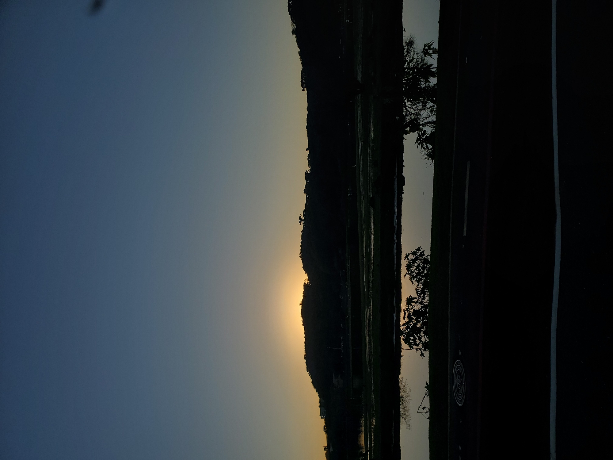 The image captures a serene twilight scene featuring a silhouetted landscape. A dark, undulating hill forms the backdrop, with scattered trees dotting its crest. Below the hill, a body of water, likely a river or lake, reflects the soft glow of the setting sun, which is partially obscured by the hillside, casting a warm yellow light. The foreground shows a dark asphalt road with white markings, and a circular manhole cover is visible on the left side of the road. The sky transitions from a deep blue at the top to a pale yellow near the horizon. The overall impression is one of tranquility and the quiet beauty of nature at dusk. The scene is not identifiable as being specifically in Curitiba, Brazil, as there are no distinguishing landmarks or characteristics to indicate this location.