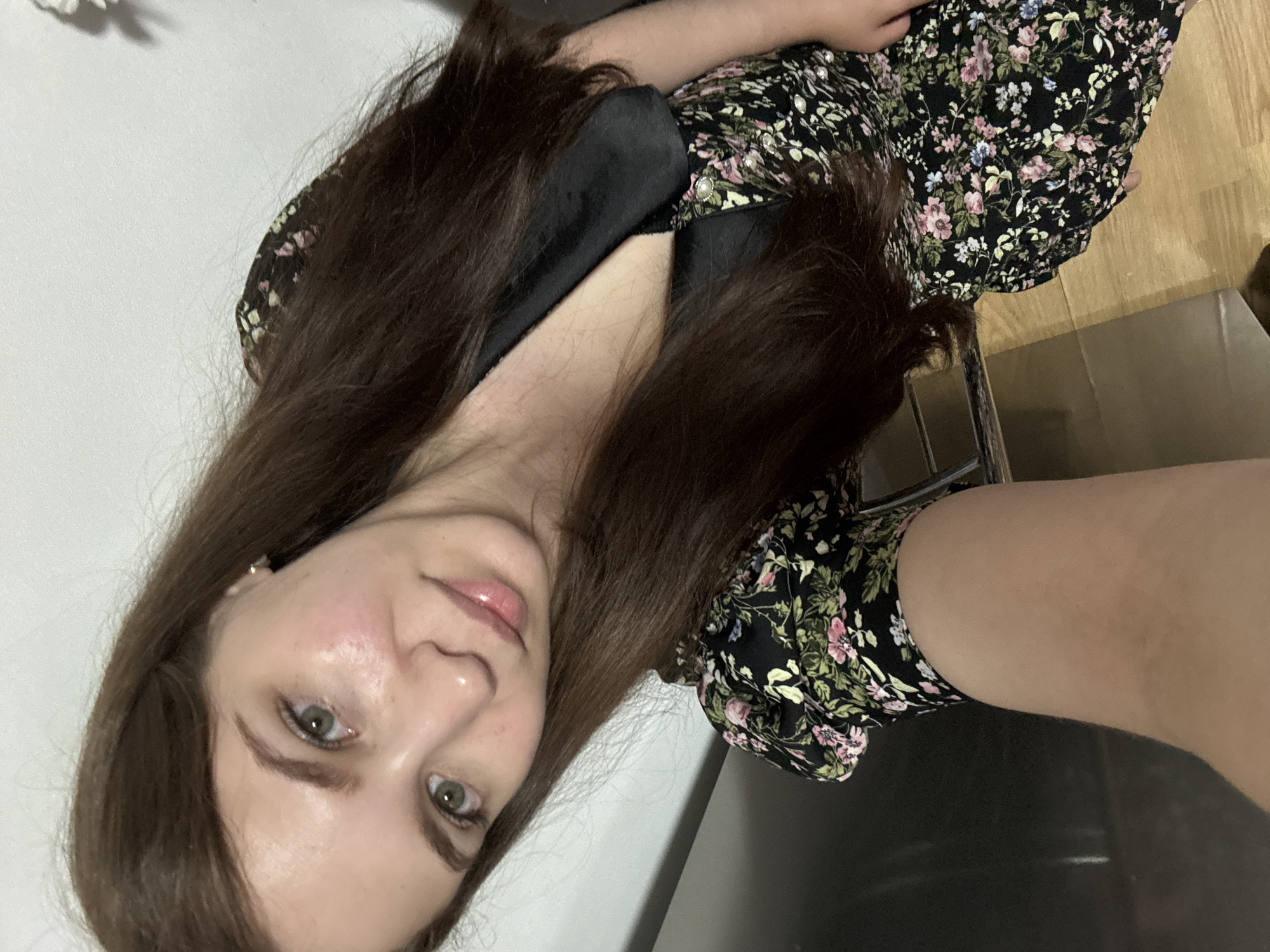 A young woman with long, dark brown hair, light green eyes, and light skin is captured in a selfie. She is wearing a black dress with a floral print featuring pink, white, and blue flowers. The dress has a black collar and pearl-like buttons. The woman is positioned with her face tilted towards the camera, her left side slightly closer. Her hair falls over her shoulders and chest. The background appears to be indoors, with a white wall on the left and a wooden floor visible at the bottom right. The overall lighting suggests an indoor setting, possibly a home environment. This image is not associated with the city of Chaikovsky, Russia.