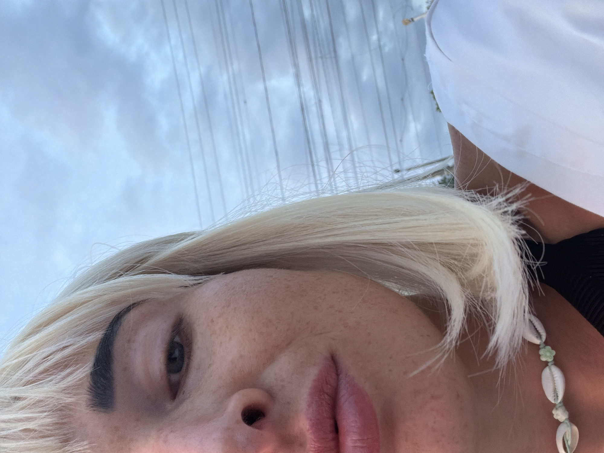 The image is a close-up shot of a person's face, with the right side of their face and blonde hair dominating the frame. The person has fair skin with freckles, dark eyebrows, and is wearing a seashell necklace. The background shows a cloudy sky with power lines stretched horizontally across it. The lighting suggests it is either early morning or late evening. The location is not specified in the image.