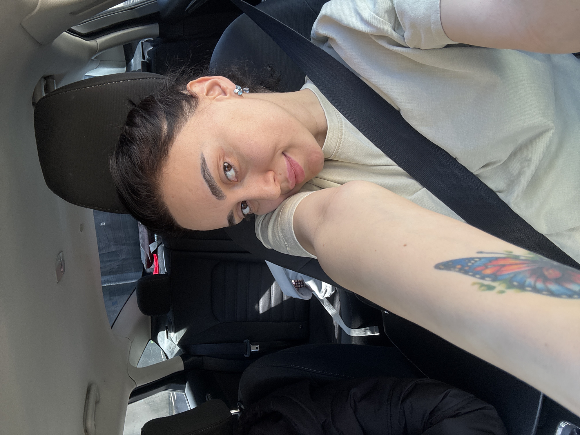 A young adult female is seated in the driver's seat of a car. She is wearing a light-colored t-shirt and a black seatbelt. She has dark hair pulled back from her face, light skin, and a small earring in her left ear. Her left arm is extended towards the camera, and a butterfly tattoo is visible on her forearm. The car's interior is black. The photo appears to be taken inside the vehicle. There is no immediate evidence to pinpoint the location as Tbilisi, Georgia.
