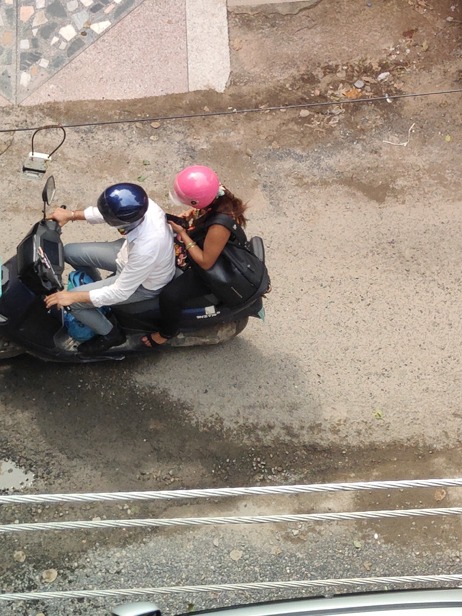 Two people, a man and a woman, are riding on a dark-colored scooter. The man is in front, wearing a white long-sleeved shirt, grey pants, a blue helmet, and black shoes. The woman is behind him, wearing a black top, black pants, a pink helmet, and black sandals. She has a black backpack on her left shoulder. They are both wearing helmets, suggesting they are in Gurgaon, India, where helmet usage is mandatory for scooter riders. The scooter is on a paved road with some debris and a wet patch, indicative of a typical urban environment in Gurgaon. In the background, there is a tiled walkway and more of the paved road.