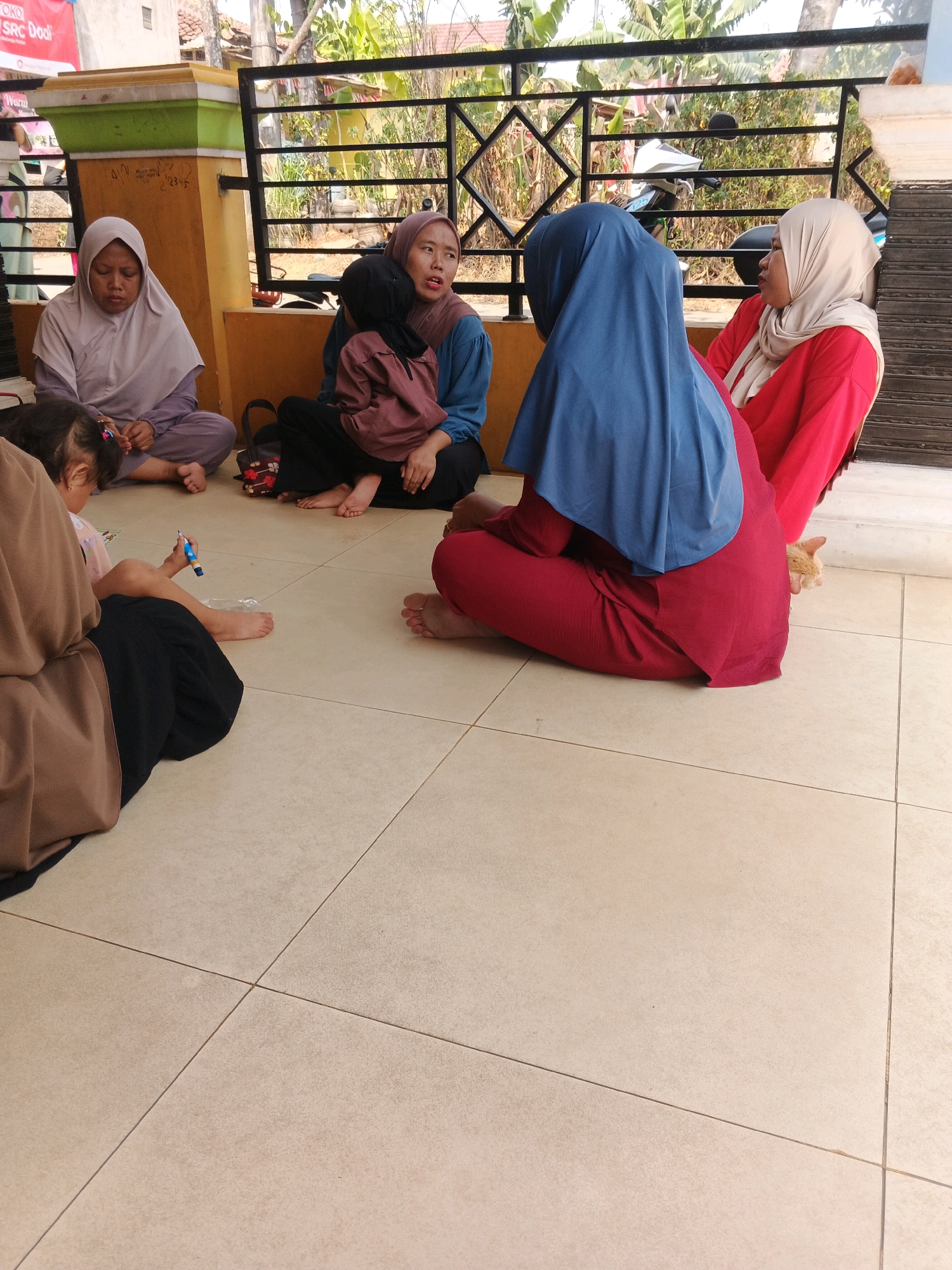 In Kadu, Indonesia, five individuals are seated on a tiled floor in front of a building with a decorative metal fence. One woman on the left wears a light purple hijab and a matching outfit, looking downwards. Next to her, a woman in a brown hijab holds a child in a dark brown outfit. Another woman, seated in the center-right, wears a blue hijab and a red outfit, facing towards the left. To her right, a woman in a light beige hijab and a red outfit is partially visible, looking away from the camera. A young child in a light pink shirt and dark pants sits in the foreground, holding a blue and yellow marker and appears to be drawing on a piece of paper or plastic. An orange cat is nestled behind the woman in the red outfit. The environment suggests a residential or community outdoor space with greenery visible through the fence.