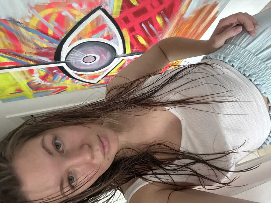 An image depicts a female individual in an indoor setting within the city of Vozdovac Urban Municipality, Serbia. The individual, appearing young to middle-aged, possesses light skin, light-colored eyes, and wet, dark brown hair hanging loosely around her face and shoulders. She is wearing a white, ribbed V-neck sleeveless top and light blue, loose-fitting bottoms with an elasticated waistband. Her left hand rests on her hip or the top of her pants. She is looking directly at the camera with a neutral expression. Behind her, a large, vibrant abstract painting dominates the background. The artwork features a prominent, stylized eye motif with a black outline, a grey/purple iris, and radiating white lines, set against a background of neon yellow, bright pink, red, blue, and orange abstract strokes. The wall behind her is white. The scene is well-lit, suggesting natural or bright artificial illumination.