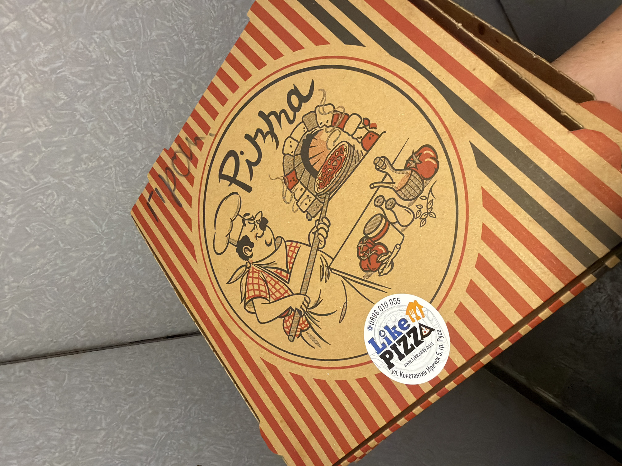 A pizza box is pictured on a textured, light gray surface, partially obscured by a dark surface. The pizza box is decorated with red and black diagonal stripes. A circular design is centered on the lid, featuring a cartoon image of a chef holding a pizza peel, a brick oven with a pizza inside, and various ingredients like tomatoes and basil. The text "Pizha" is written in a stylized font above the oven. A circular sticker is placed on the left side of the design, with the logo "Like PIZZA". Below the logo, text in Cyrillic and Latin script reads "ул. Константин Иречек 5, гр. Русе" (ul. Konstantin Irechek 5, gr. Ruse), indicating the address is located in Ruse, Bulgaria. Additional text on the sticker includes a phone number and a website address.