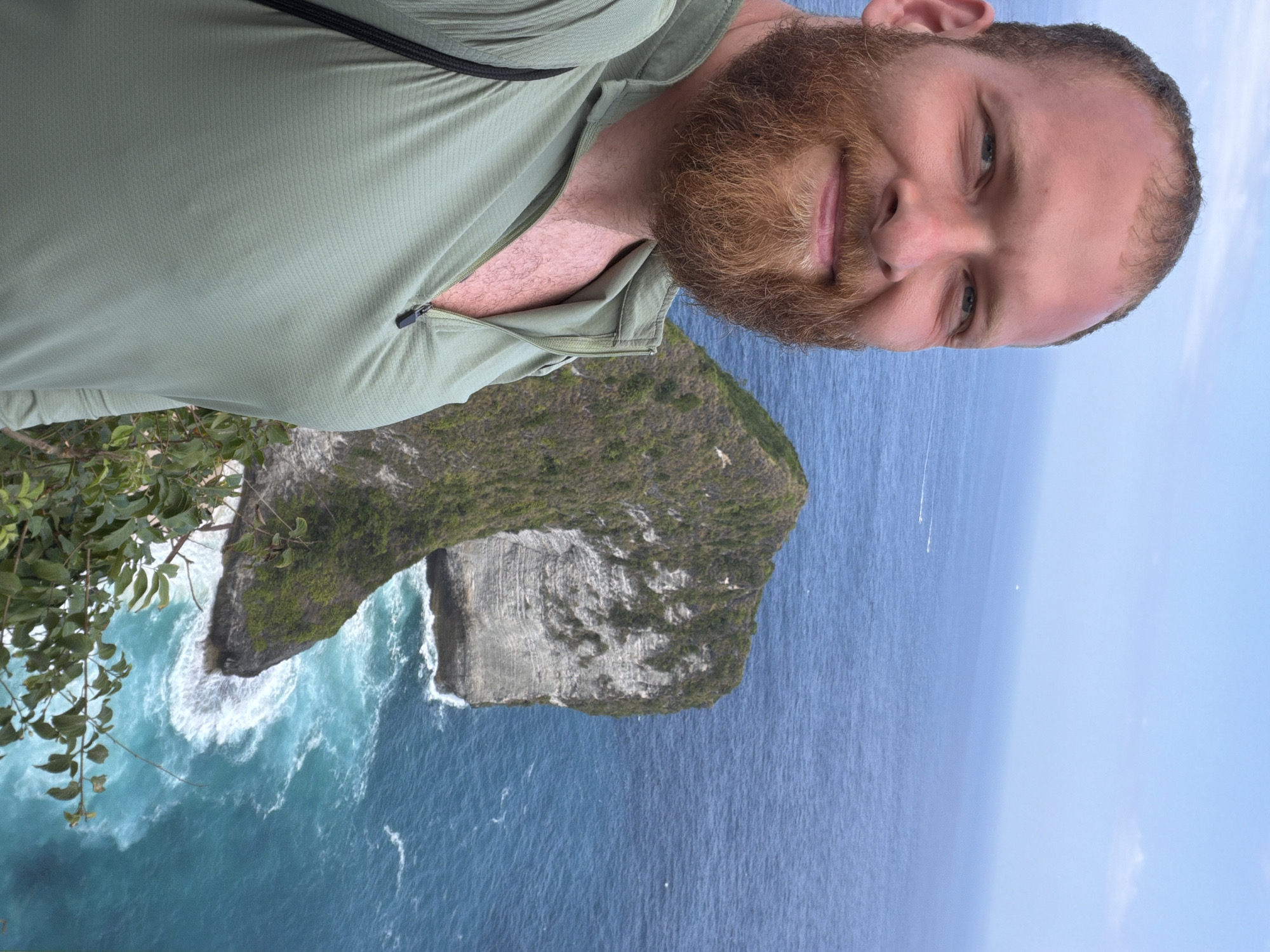 A man with a beard is taking a selfie on a cliff overlooking the ocean. He is wearing a light green, zip-up shirt. In the background, a prominent rock formation juts out of the turquoise water. The sky is clear and blue, with a few white clouds. The scene appears to be from Nusa Penida, Indonesia, likely at a scenic viewpoint such as Kelingking Beach.