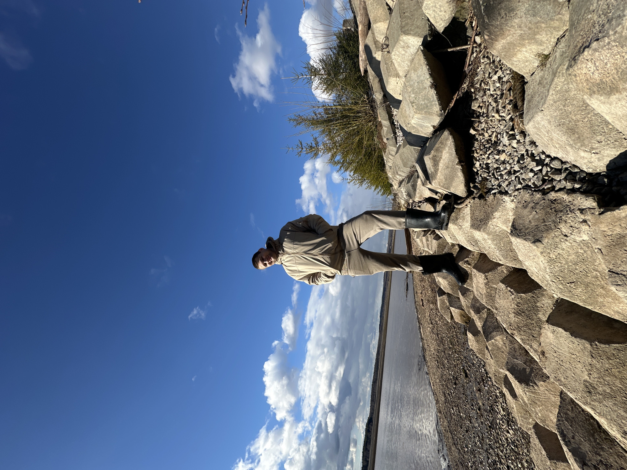 A man stands on a structure made of concrete blocks, likely a breakwater or dam, next to a body of water under a blue sky with scattered clouds. The man is wearing a light-colored jacket and pants, and dark boots. He is facing the camera. Behind him is a mix of gravel and more concrete blocks. The location is likely in Посёр, Russia, given the prompt. The water appears to be a river or lake, with trees visible on the opposite shore in the distance. The sun is shining, casting shadows on the concrete structure.