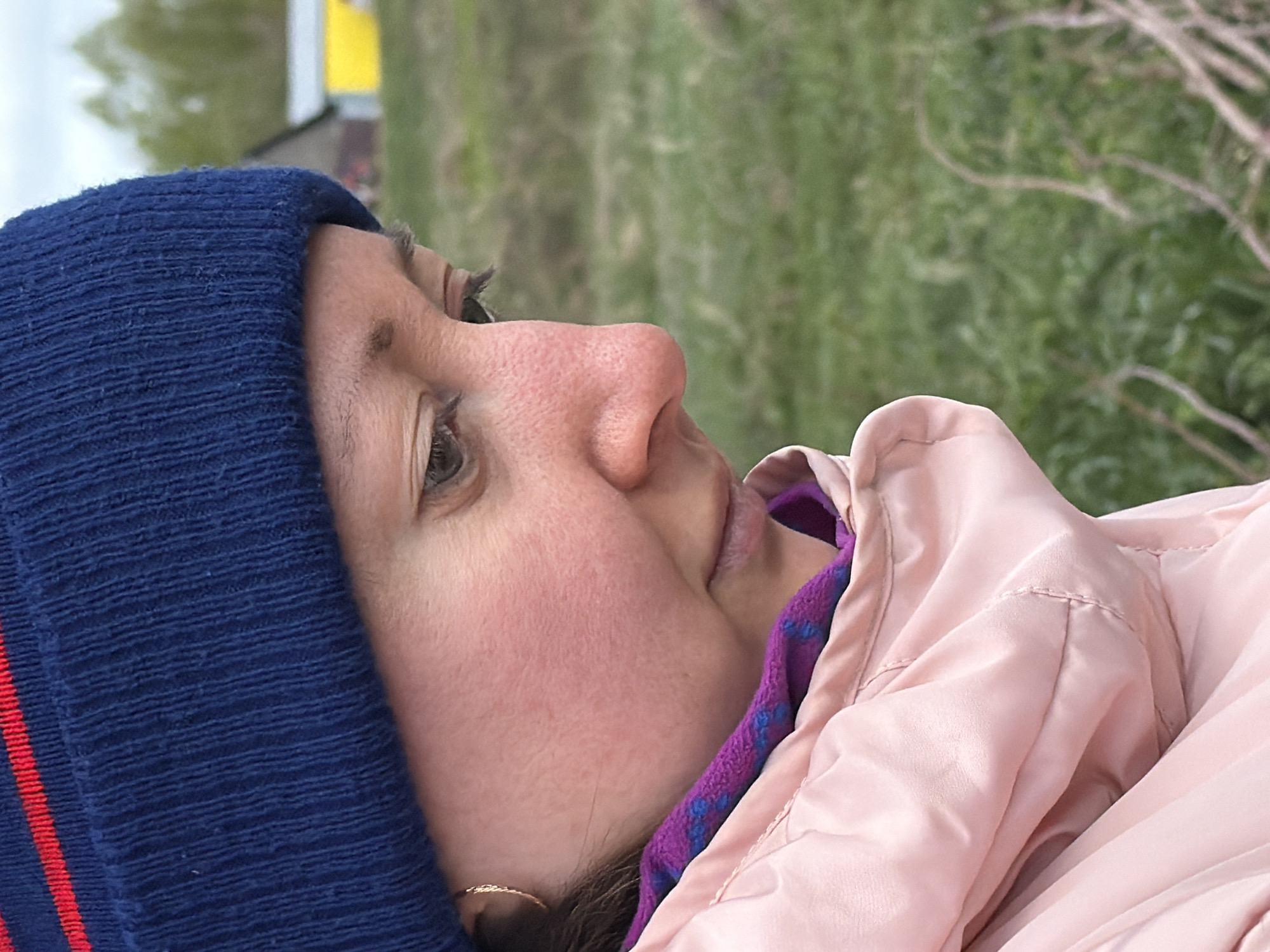 A close-up, rotated, outdoor photograph shows a person wearing a navy blue knit hat with a red stripe, a pink jacket with a purple and blue patterned lining, and a gold hoop earring. The person's head is oriented to the left of the frame. The background consists of green vegetation and a blurred building. The photograph may have been taken in Ol'khovo, Russia.