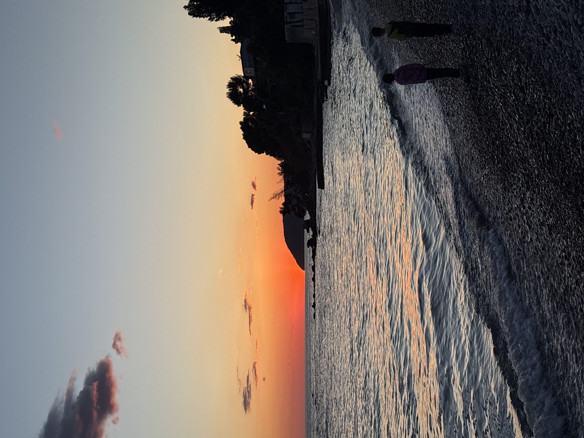The image displays a serene coastal landscape at what appears to be sunset. The sky, dominating the upper portion, transitions from a vivid orange and red at the horizon, indicative of the setting sun, to softer yellow and light blue-grey tones higher up. A few small, dark clouds with hints of pink are visible in the upper-left sky.
A body of water, likely the sea, stretches across the middle of the frame, with gentle waves breaking onto a dark, pebbly beach in the foreground. The water's surface beautifully reflects the warm colors of the sky, creating shimmering streaks of orange and yellow amidst the ripples.
Two individuals stand on the pebbly shore, close to the water's edge. Their forms are silhouetted against the water and sky. One person wears a light-colored top (possibly yellow-green) and dark trousers, while the other wears a darker top (perhaps purple or dark blue) and dark trousers. They appear to be observing the sea or the sunset in a contemplative manner.
On the left side, a dark, silhouetted landmass, covered with trees and some structures, outlines the coastline, suggesting hills or cliffs. The textures of the water's ripples and the pebbles on the beach are distinctly visible. The scene evokes a tranquil atmosphere, characteristic of a scenic coastal area such as Bar, Montenegro. No visible text is present.