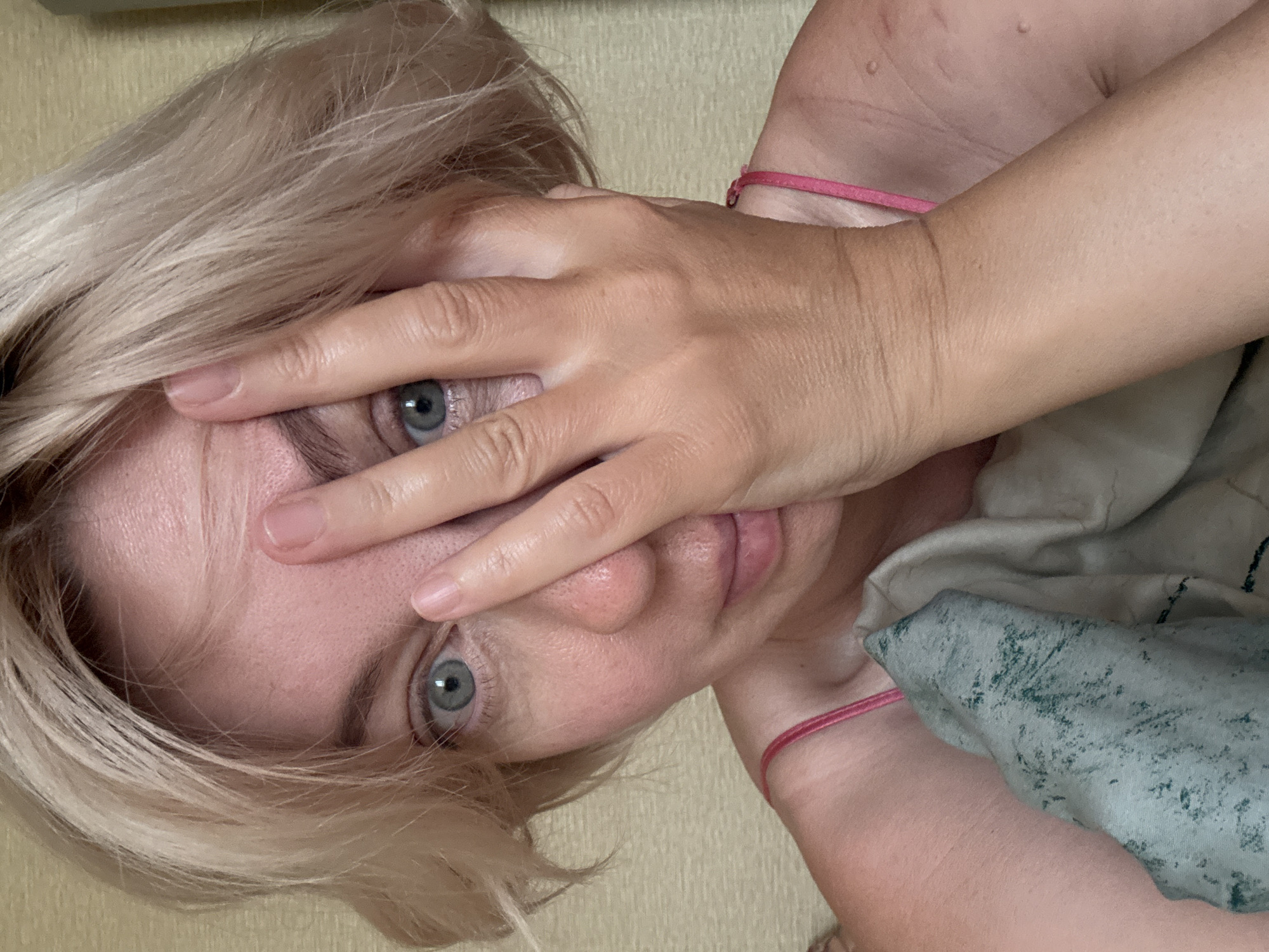 A woman with short blonde hair is partially covering her face with her right hand, revealing one open eye. She is wearing a pink camisole. The background is a textured beige wall. The image appears to be a selfie taken indoors, possibly in a residential setting in Sochi, Russia. There is a blanket or sheet in the lower part of the frame.