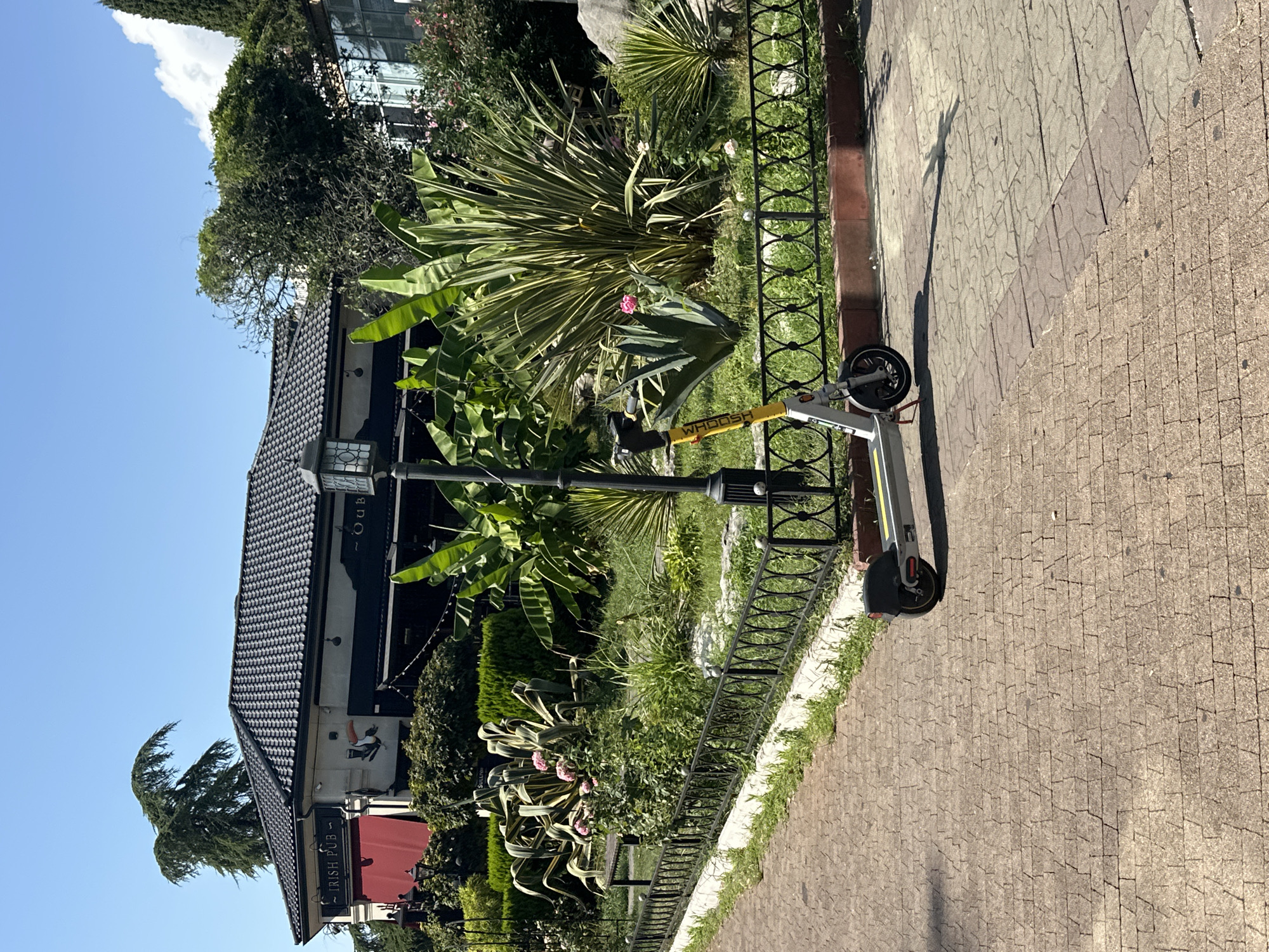 In Sochi, Russia, an outdoor scene features a light-colored electric scooter with yellow accents parked on a paved walkway. The scooter is positioned in the foreground to the right of a black metal fence. Beyond the fence, a lush green garden is visible, with various plants, including large-leafed plants and pink roses. A building with a dark tiled roof and a red awning, marked "IRISH PUB," stands behind the garden. The sky is clear and blue, indicating a sunny day. A lamppost stands near the scooter.