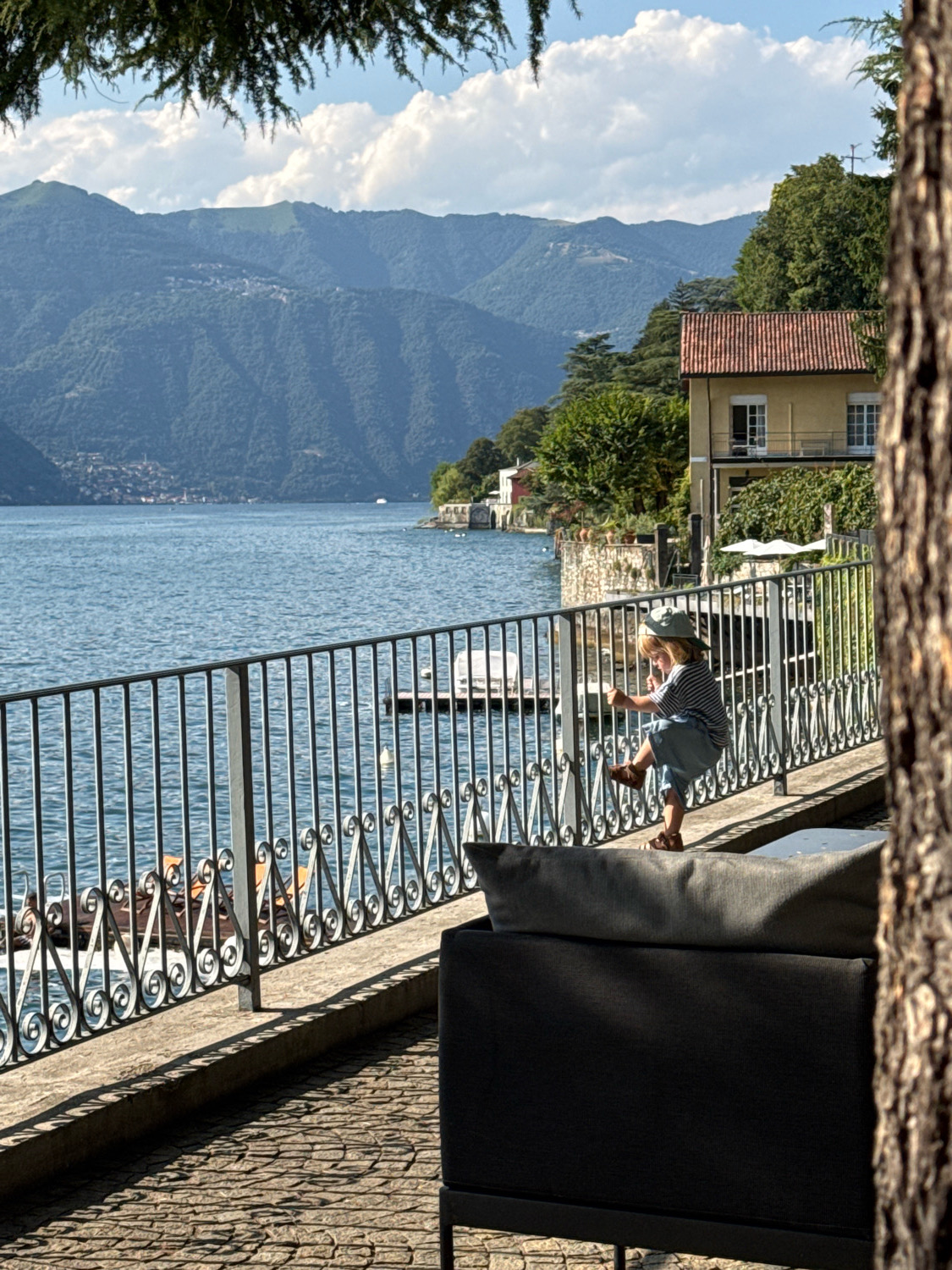 A young child wearing a striped shirt and blue overalls, with a hat on, stands on a railing, facing the water. The child's left leg is extended, and their right leg is bent. The child is on a paved walkway beside a large body of water, likely Lake Como, with a row of boats visible on the water. In the background, lush green mountains rise, with scattered buildings on their slopes. To the right of the child, a portion of a tree trunk is visible, obscuring part of the view. A dark upholstered sofa is positioned in the foreground, to the right of the frame. The scene is bathed in bright sunlight, casting shadows on the walkway. The child appears to be in Pognana Lario, Italy, given the context of the image.