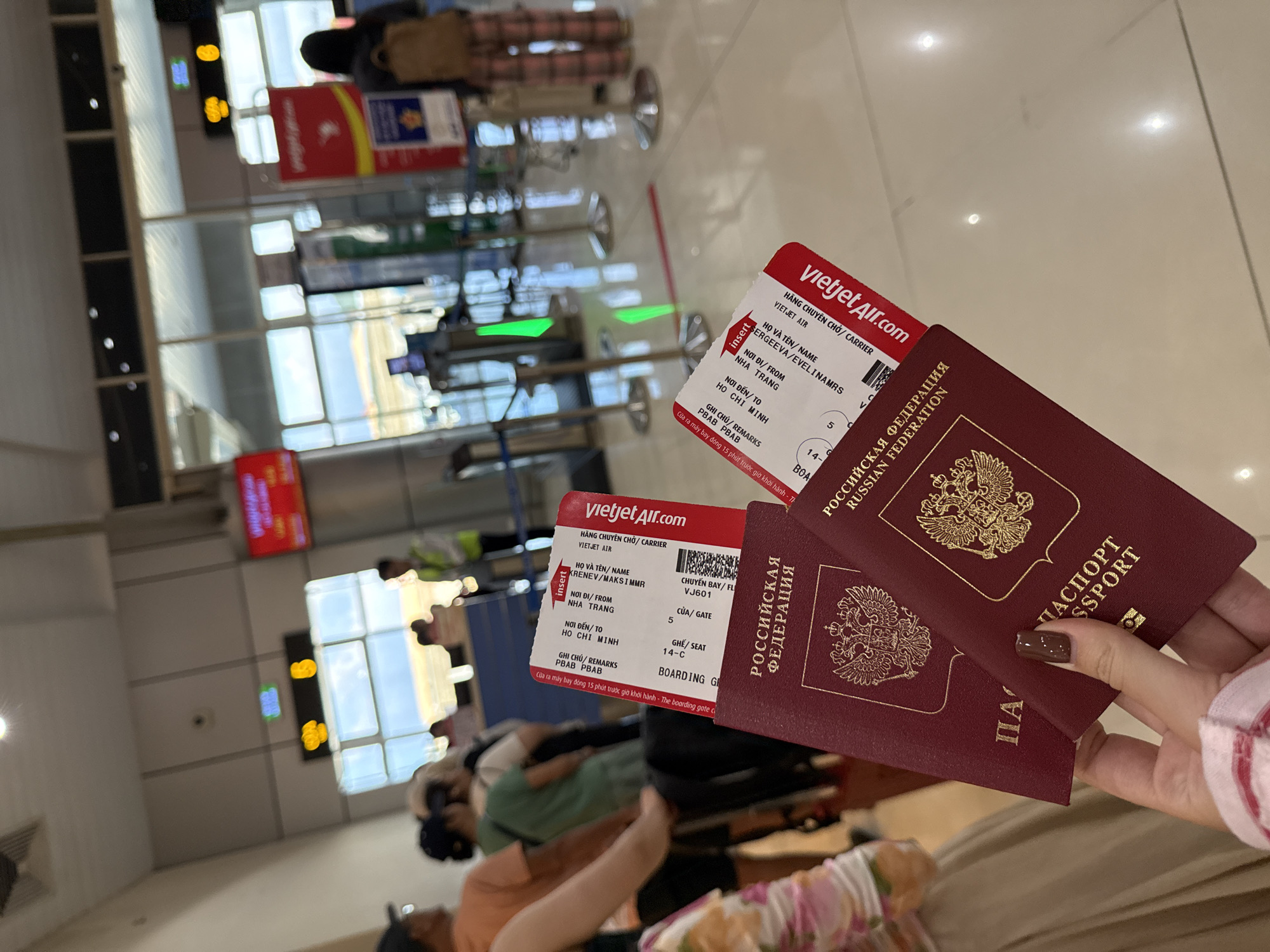 A person is holding two Russian passports and two VietJet Air boarding passes inside an airport in Bac Cam Ranh Ward, Vietnam. One boarding pass is for SERGEEVA/EVELINAMRS and the other for KRENEV/MAKSIMMR. Both boarding passes are for a flight from Nha Trang to Ho Chi Minh on flight VJ601, gate 5, seat 14-C.