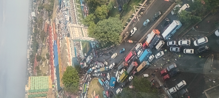 Aerial view of a congested urban intersection. Numerous vehicles, including cars and buses, are tightly packed, indicating a traffic jam. Pedestrians are visible amidst the vehicles. The scene is surrounded by buildings, trees, and roadways. The presence of emergency personnel suggests an incident or accident. A bridge crosses over the intersection.
